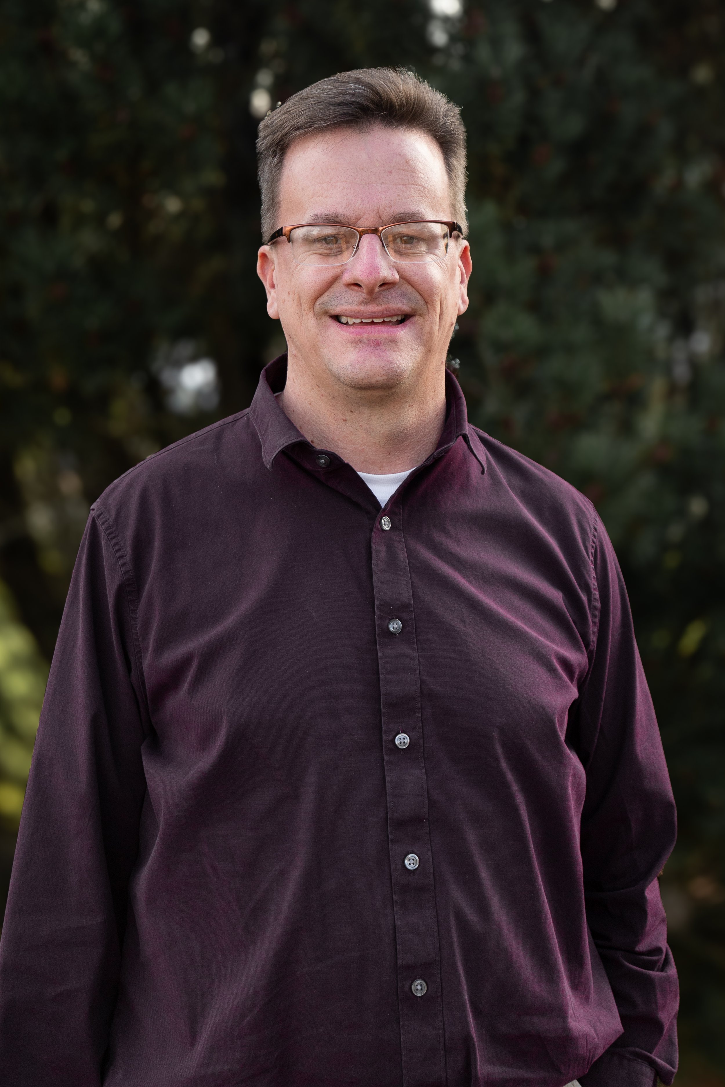 A man with glasses and short brown hair wearing a dark purple button-up shirt standing outdoors in front of trees.