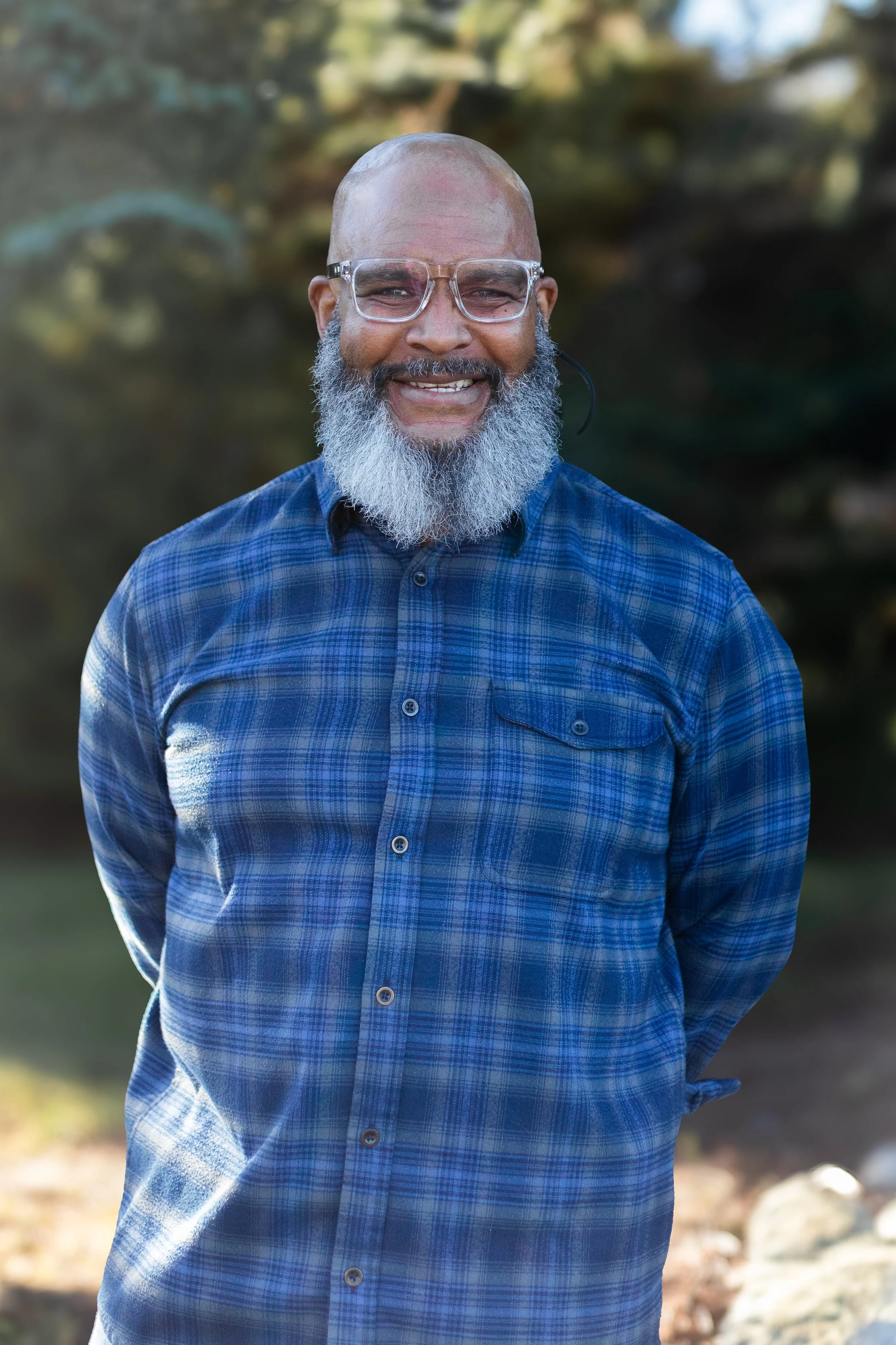 A smiling man with a gray beard, glasses, and a bald head wearing a blue plaid shirt outdoors with blurred trees in the background.