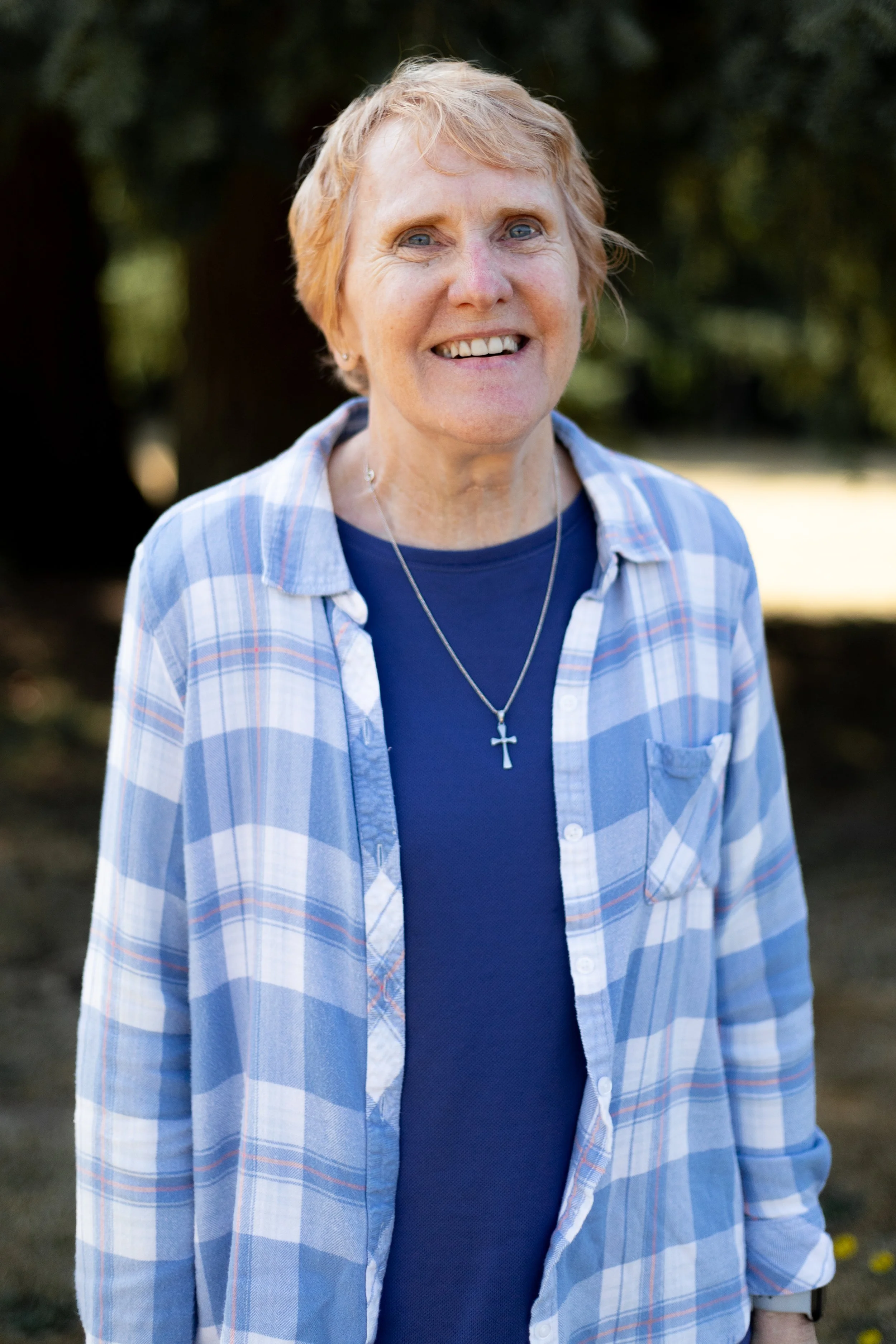 A smiling senior woman with short blonde hair outdoors, wearing a blue plaid shirt over a dark blue top and a cross necklace.