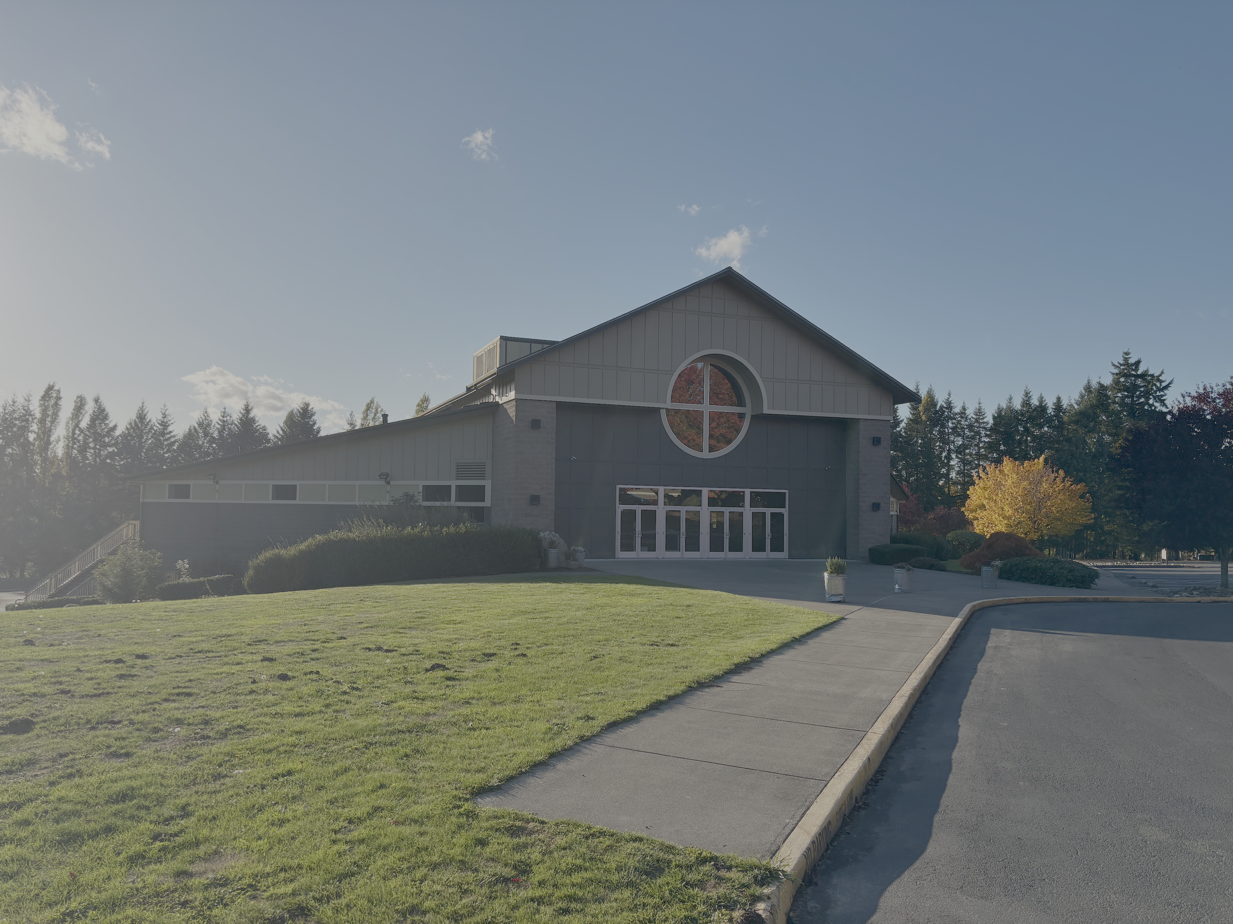 Modern church building with large circular stained glass window, surrounded by a lawn and trees with fall foliage, paved entrance, and parking lot.