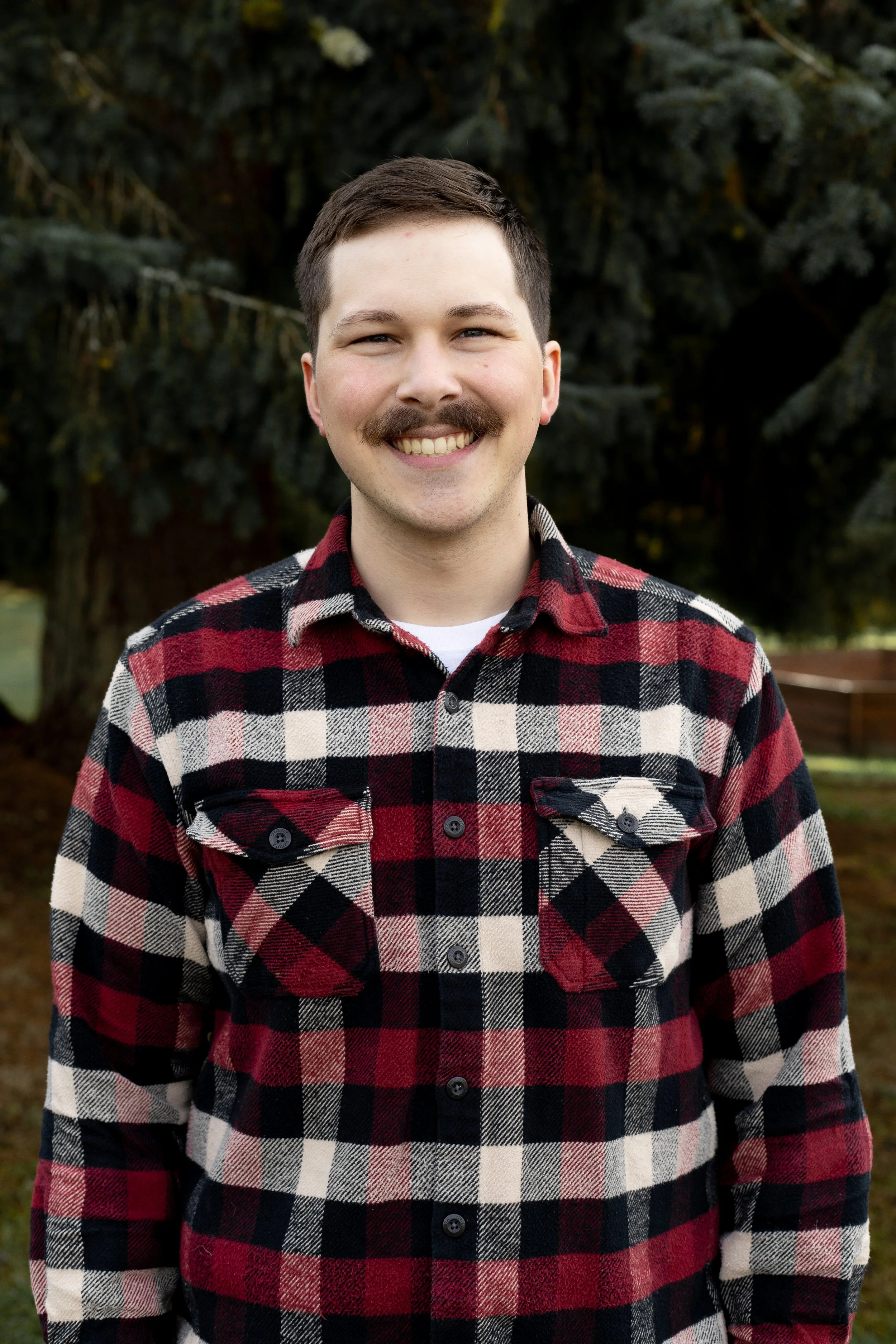 A smiling man with a mustache wearing a red, black, and white plaid shirt outdoors near trees.
