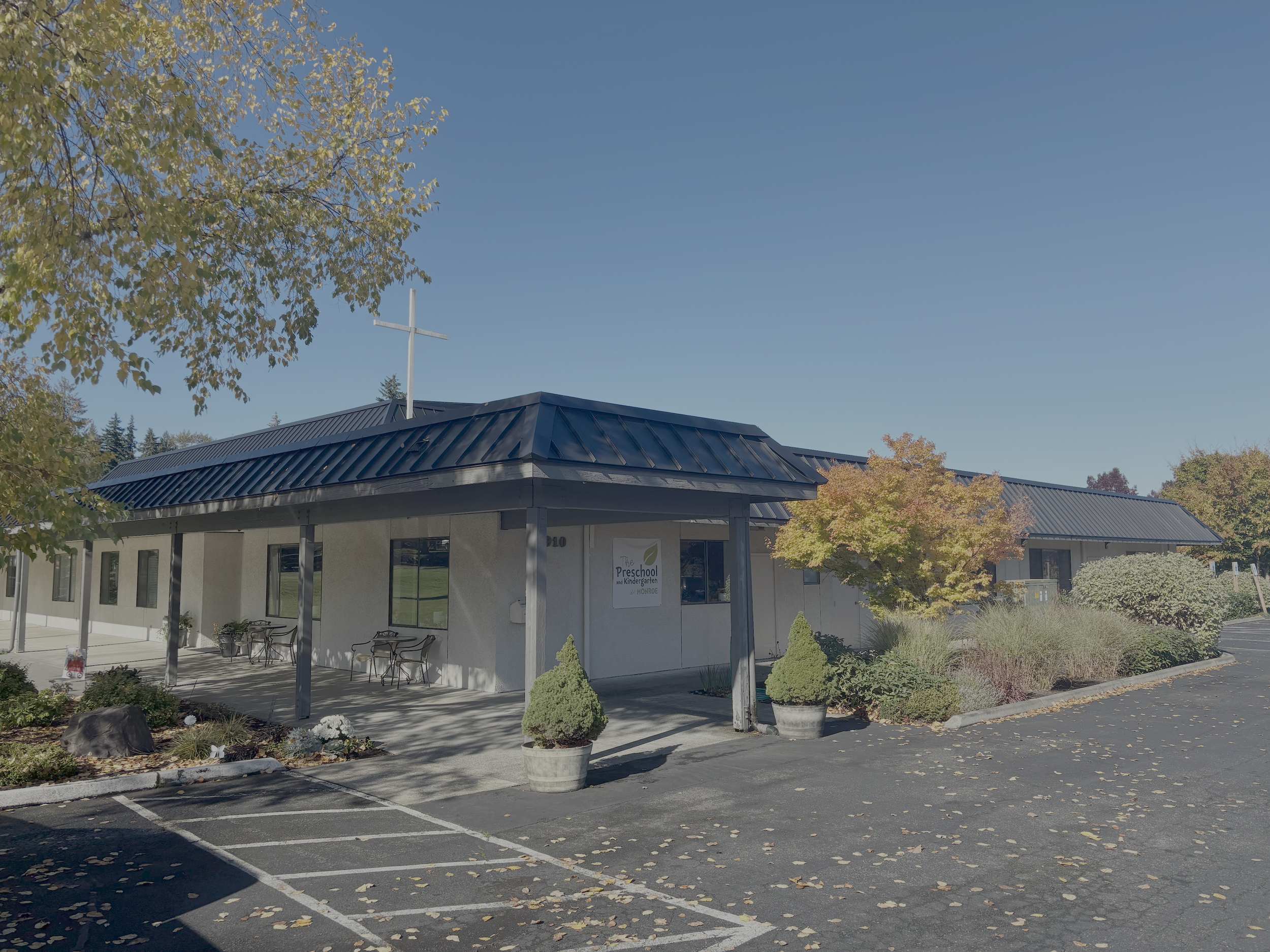 A single-story preschool building with a metal roof and outdoor seating area, surrounded by fall foliage and parking lot.