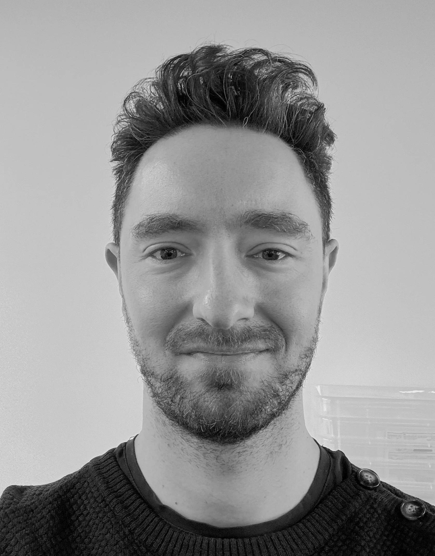 Sort-hvid portrætfoto af en ung mand med mørkt, krøllet hår og skæg, iført en mørk sweater med knapper på skulderen, smilende mod kameraet.