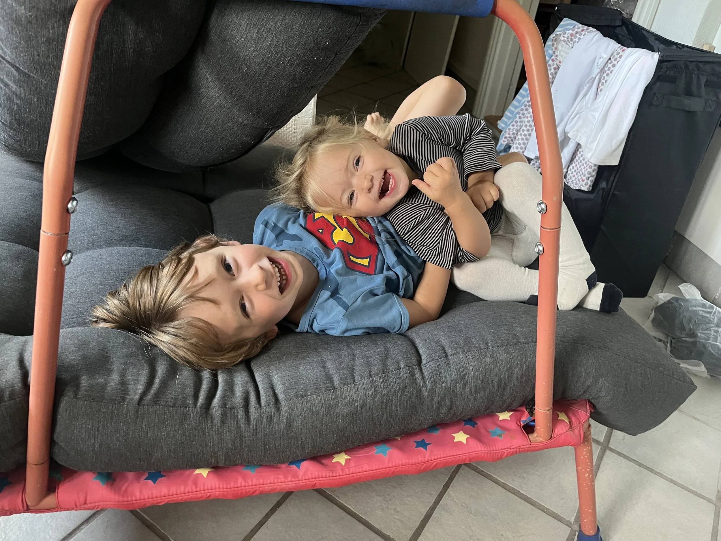 To børn leger på en sofa, hvor den ene ligger med hovedet mod kameraet, og den anden sidder på den andens mave. Begge smiler og ser glade ud.