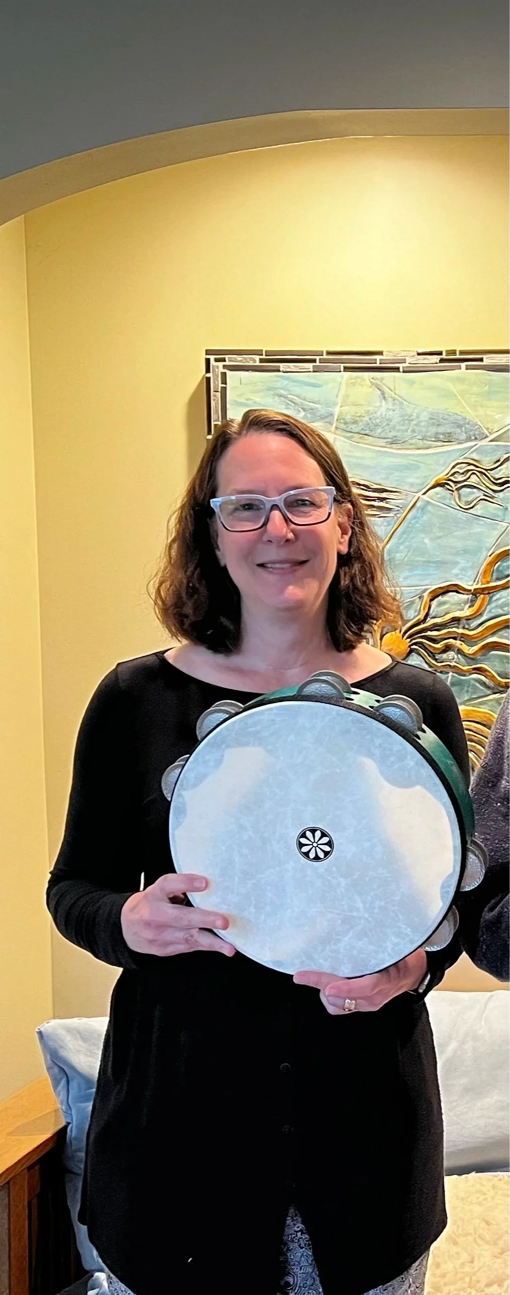 Woman with glasses holding a tambourine, standing in a room with yellow wall and colorful artwork behind her.