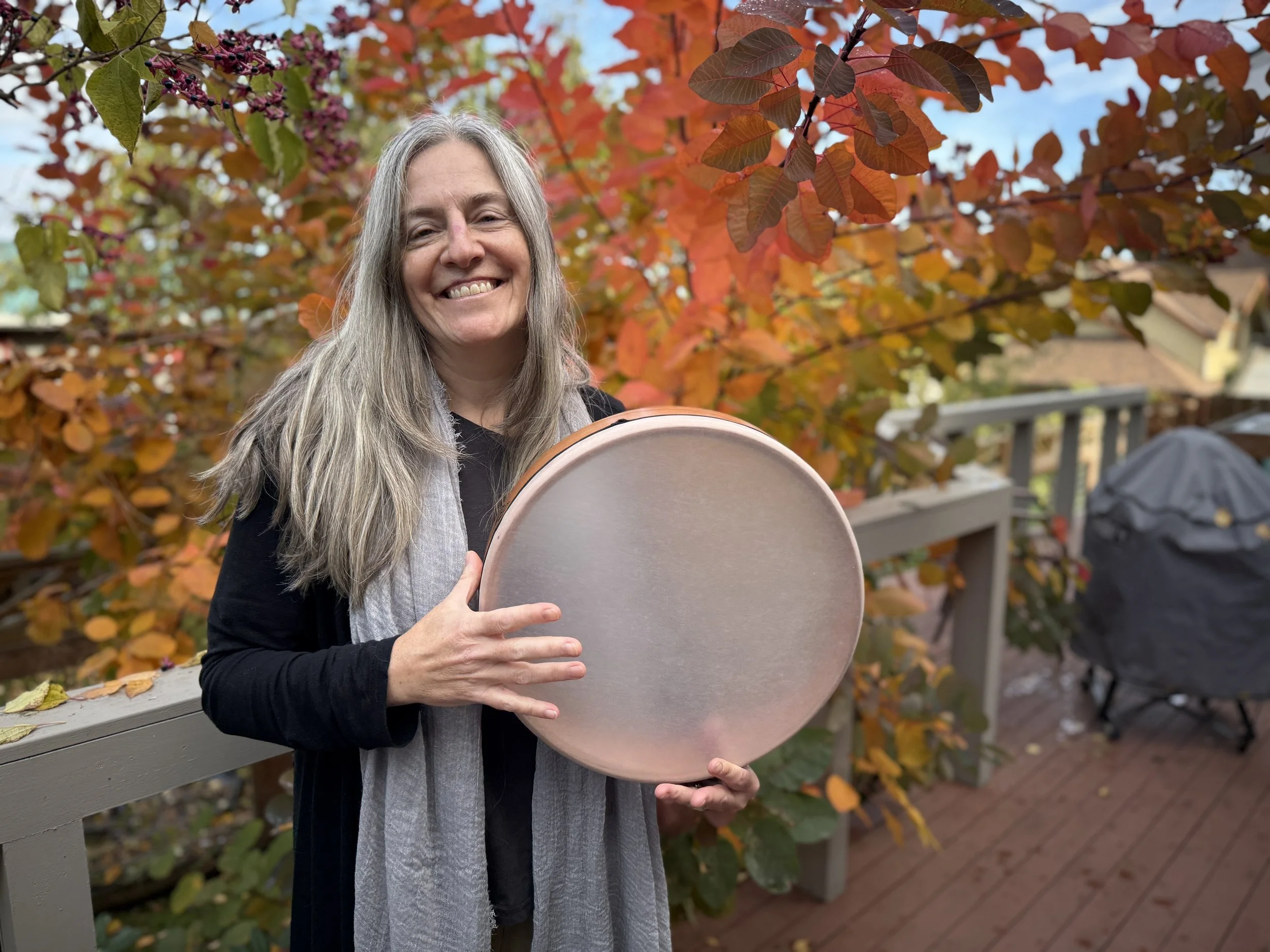 A smiling woman with long gray hair holding a frame drum outdoors on a wooden deck, with fall foliage in the background.