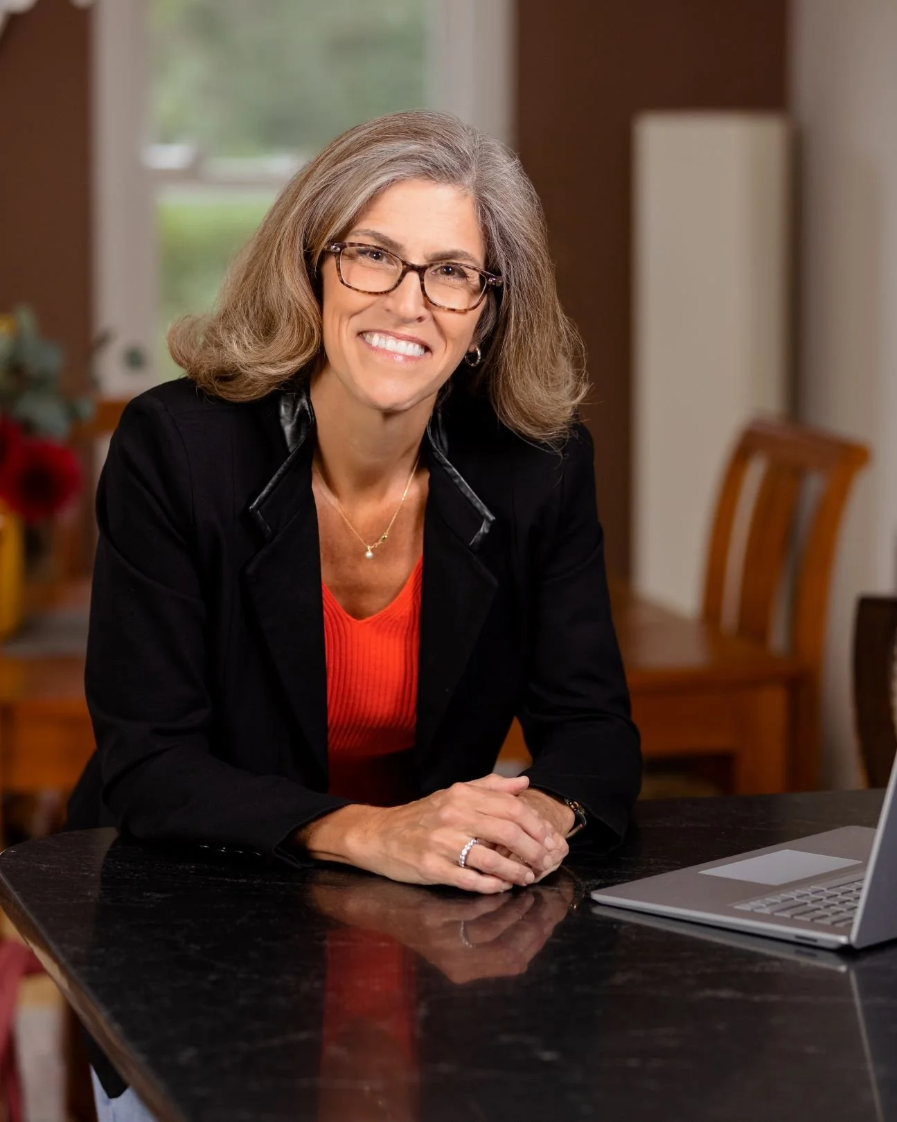 A smiling woman with gray hair and glasses sitting at a table with a laptop, in a home setting with a window and chairs in the background.