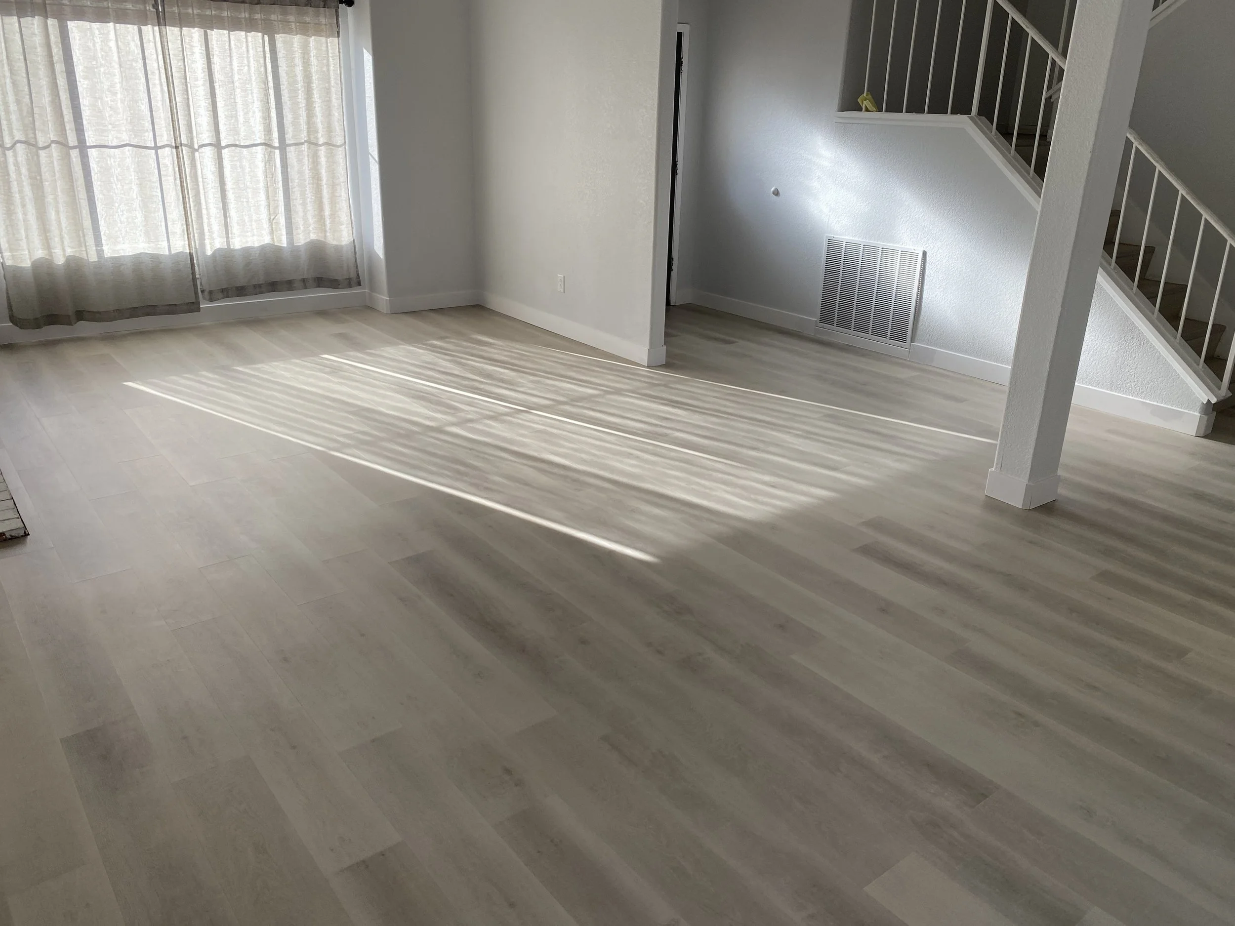 Empty living room with light-colored wood flooring, sunlight streaming through curtains, stairs with a white railing, and a wall vent.