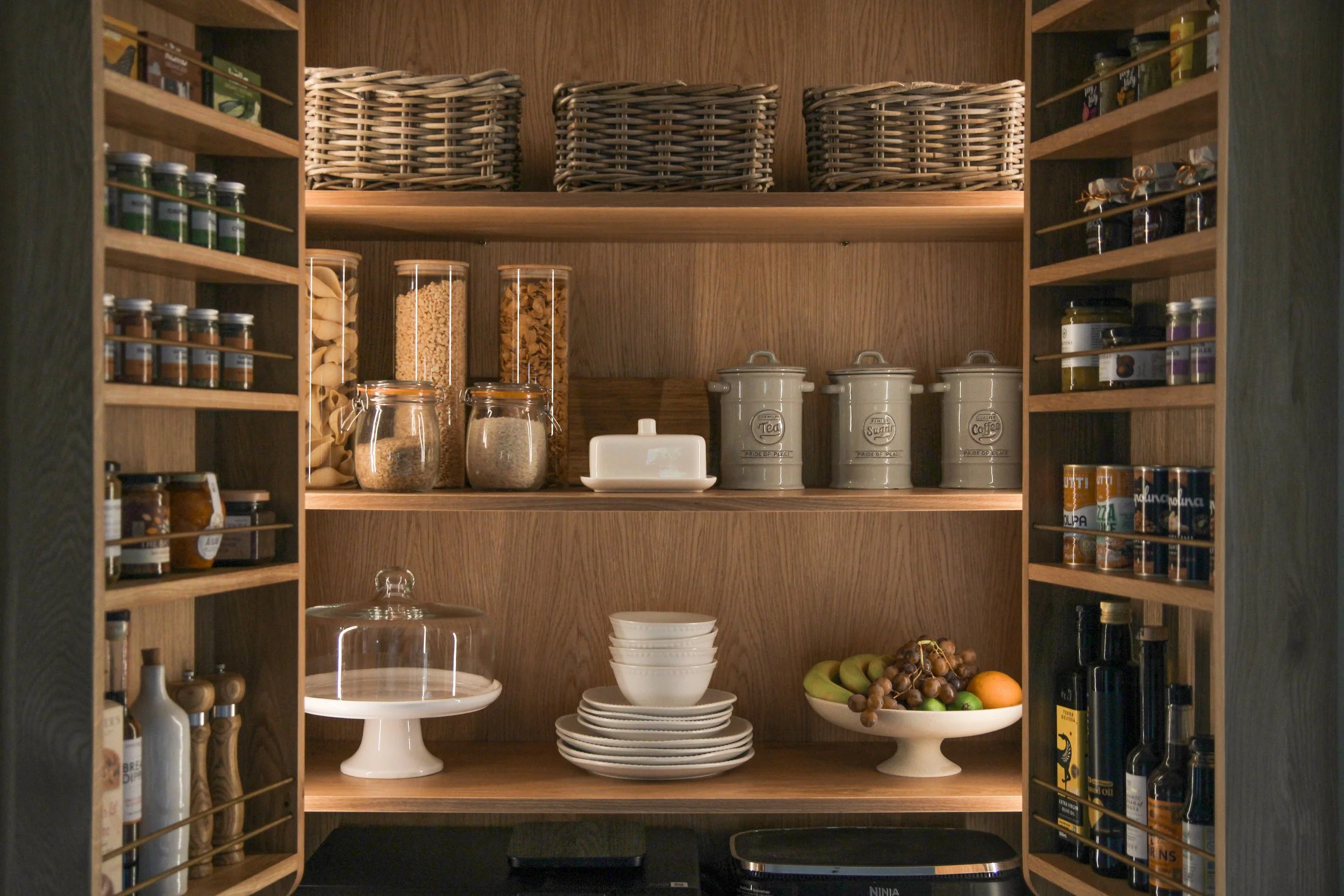 A oak kitchen double pantry cabinet filled with jars and cans of food, bowls, and decorative baskets.