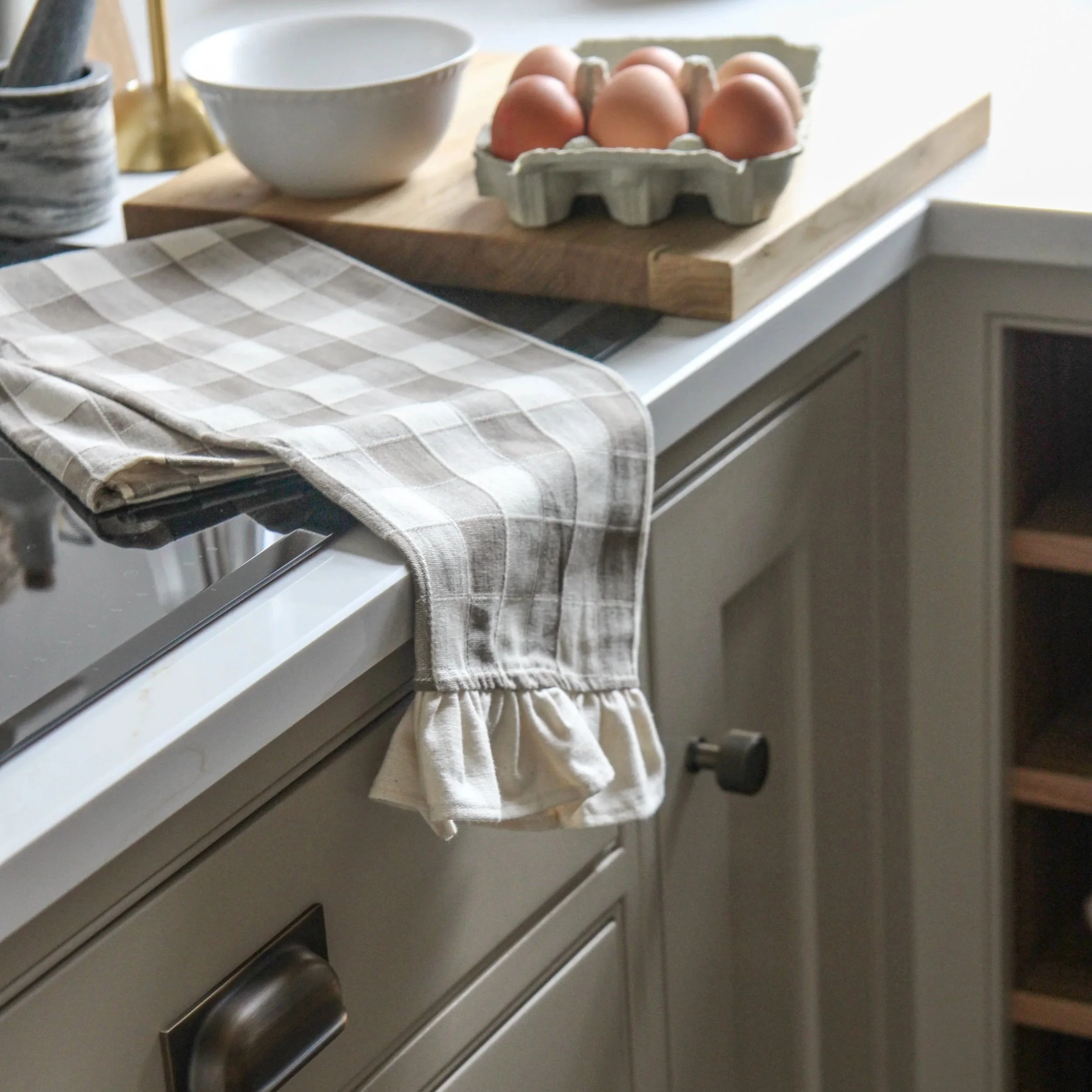 Kitchen countertop with a checkered dish towel hanging over the stove edge, a carton of eggs, a white bowl, and a wooden cutting board.