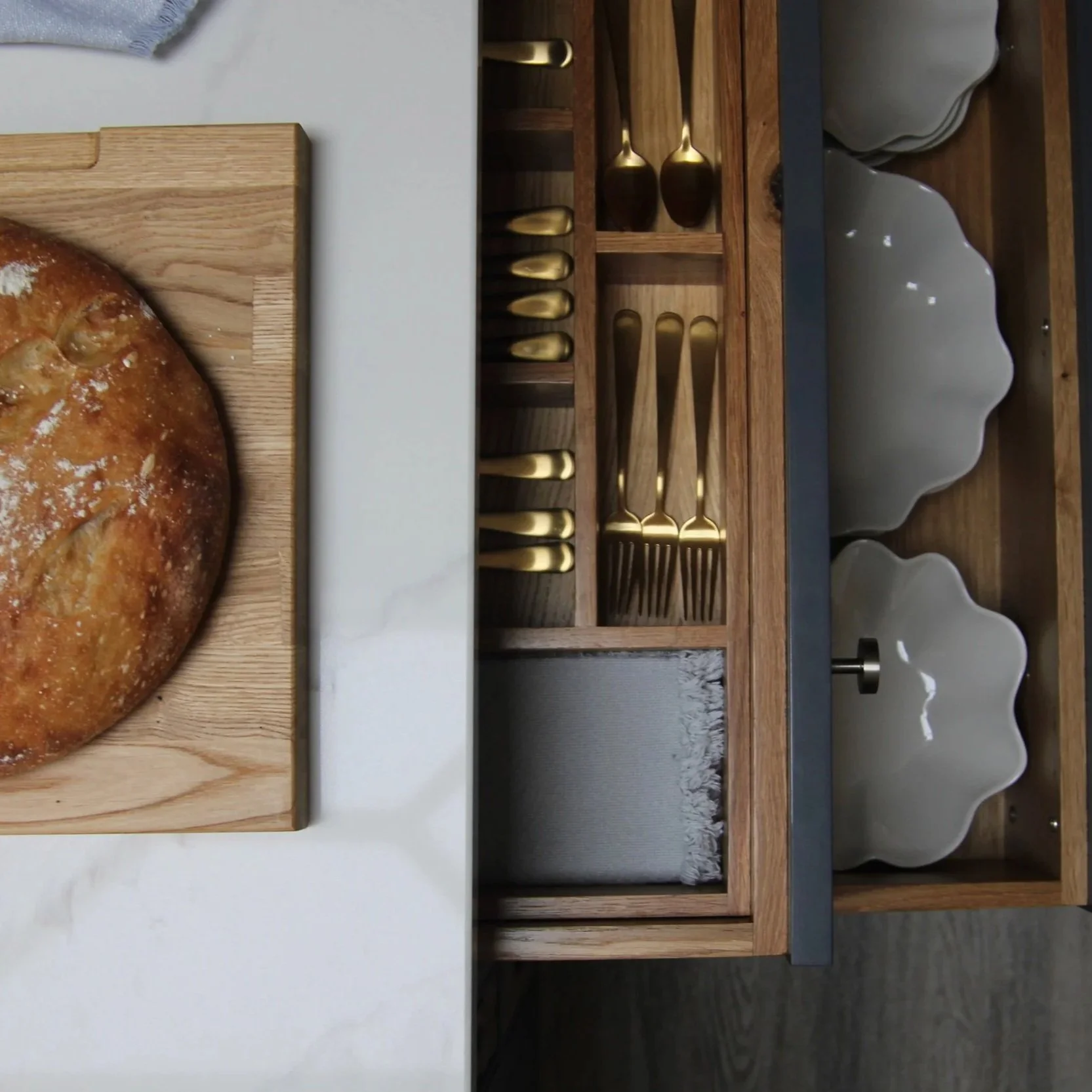 A wooden knife block with gold utensils, next to white scalloped bowls in a kitchen drawer, and a loaf of bread on a wooden cutting board.