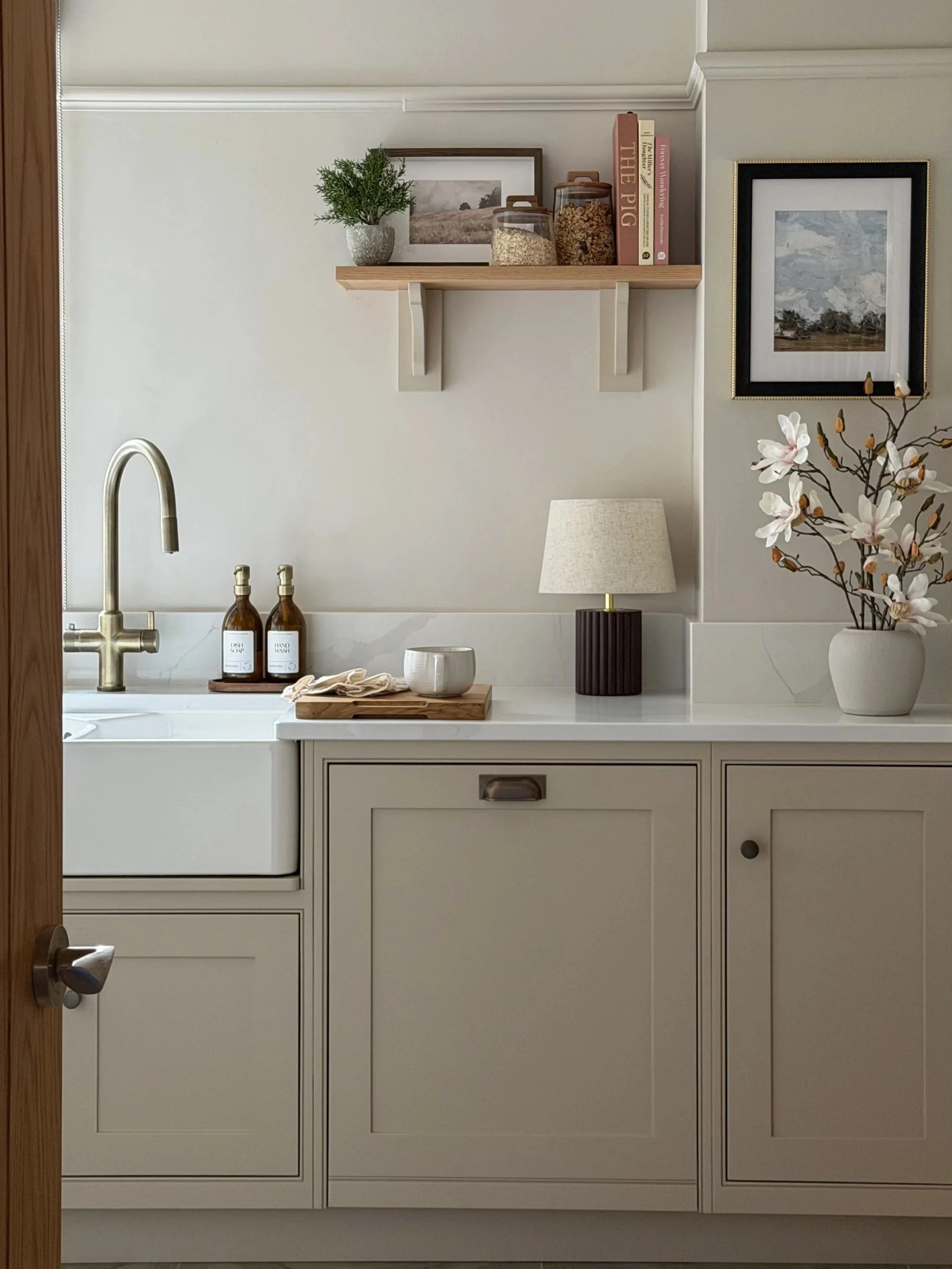 Modern kitchen with neutral shaker style cabinets, a white Belfast sink with a antique brass tap and decorative items including soap dispensers, a small lamp, a plant, framed pictures, and a vase with flowers.