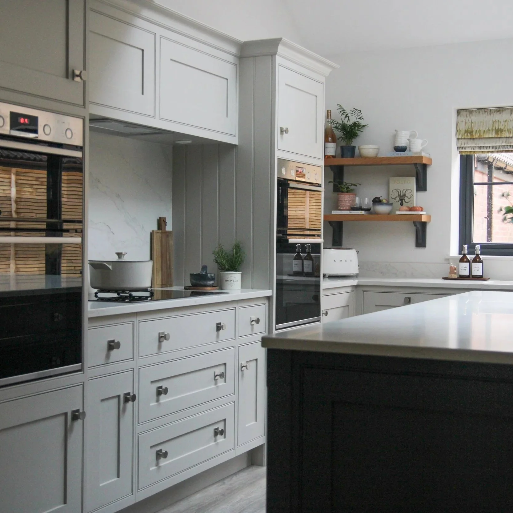 Modern shaker style kitchen with neutral cabinetry, black double oven, marble backsplash, open wooden shelves with plants and dishes, window with roman shade, and a contrasting kitchen island in foreground.
