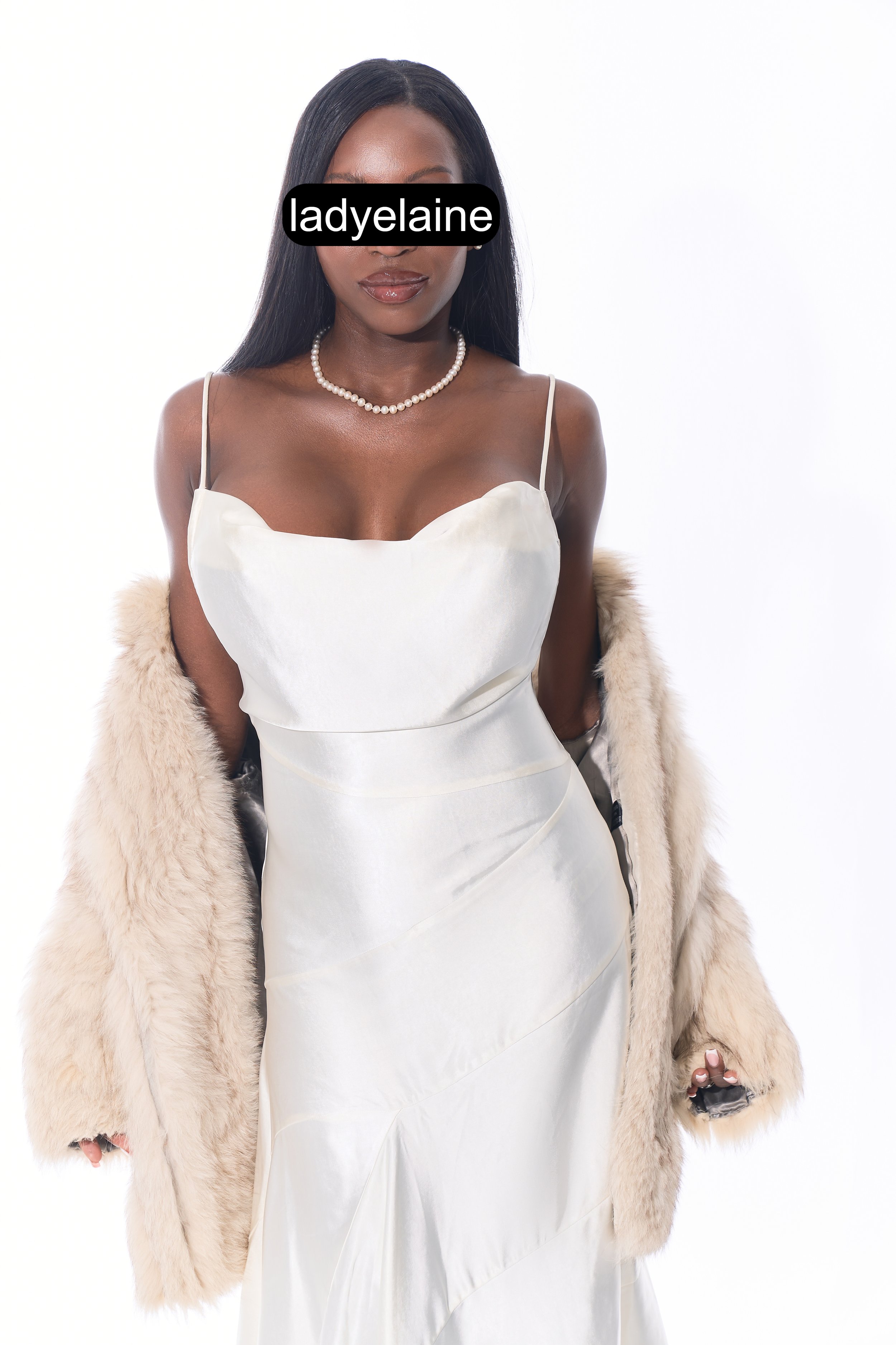 A woman with long black hair wearing a white satin dress with spaghetti straps, a pearl necklace, and a beige fur coat draped over her shoulders against a plain white background.