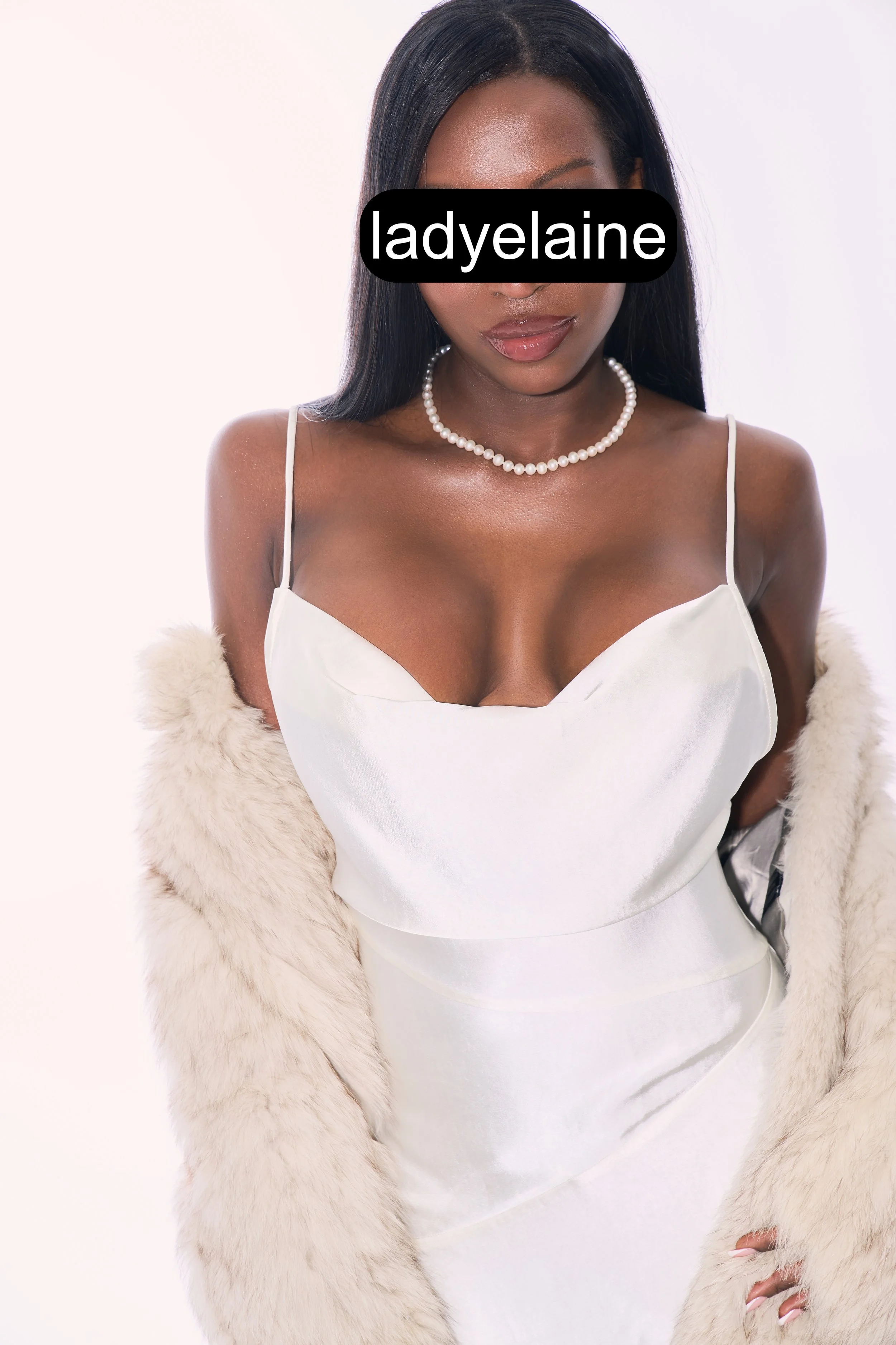 A woman with long dark hair wearing a white satin dress, a pearl necklace, and a cream-colored fur coat draped over her shoulders.