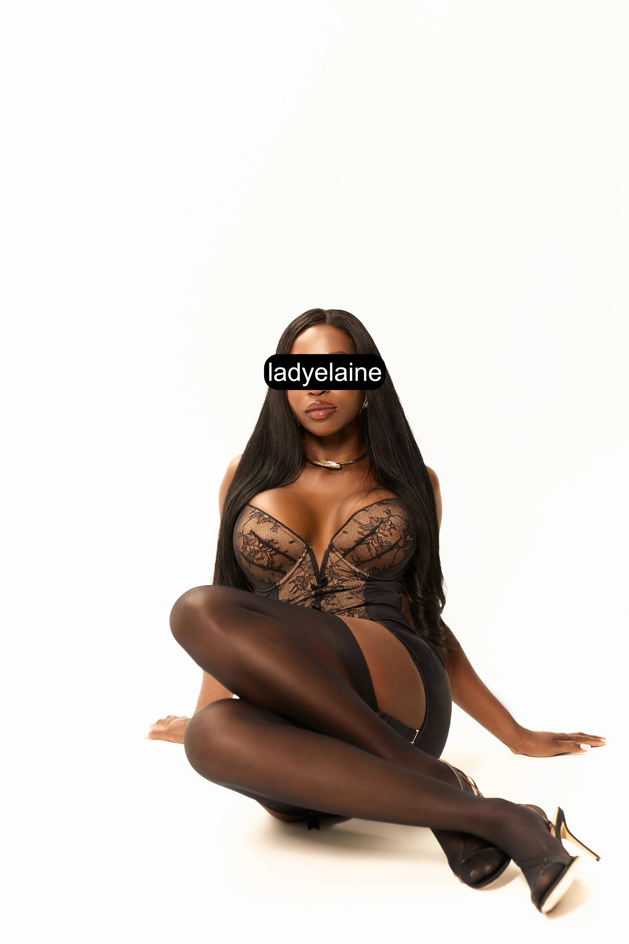 A woman in black lingerie and stockings sitting on a white background with her legs crossed.