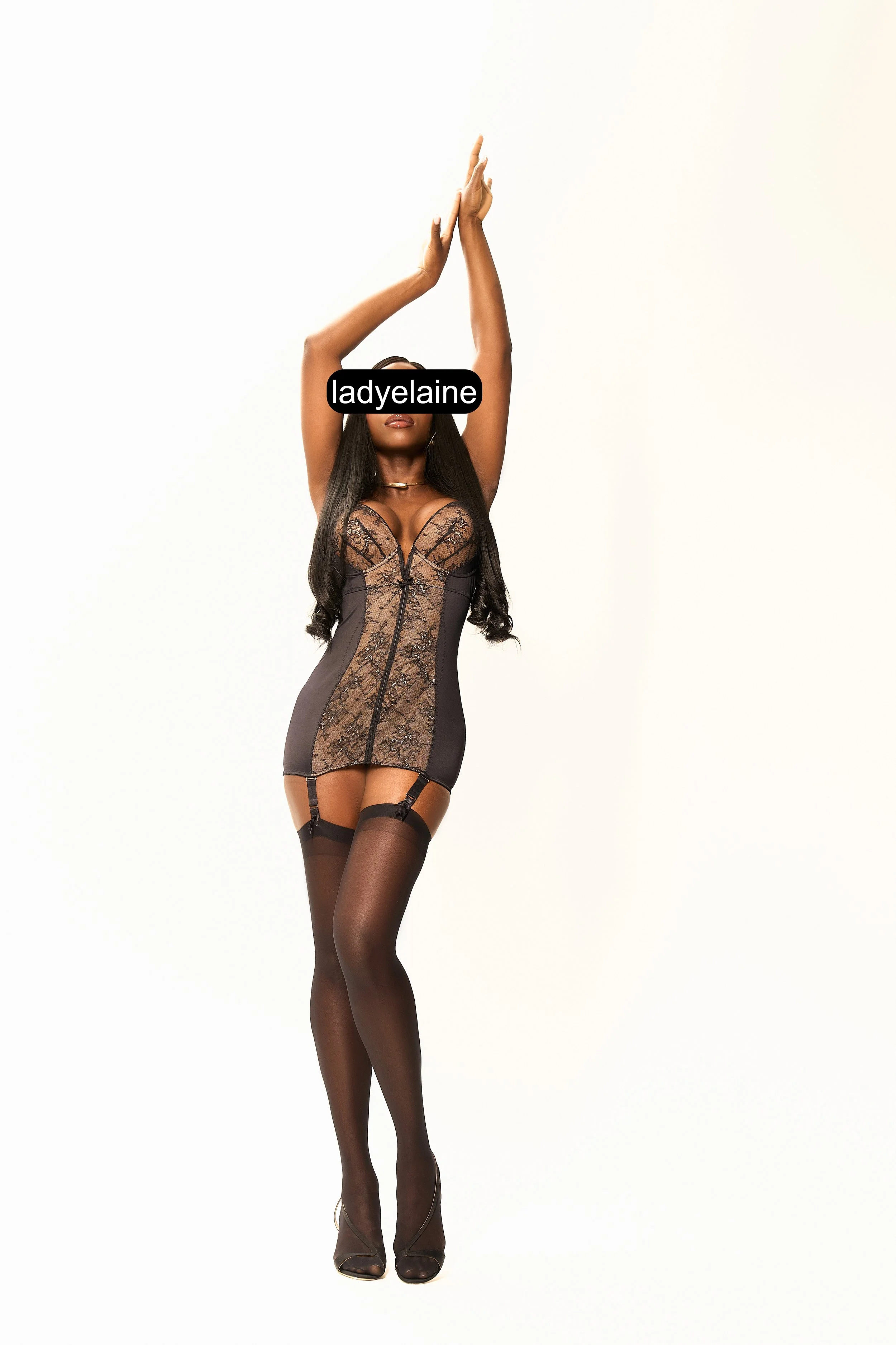 Woman in black lace bodysuit and thigh-high stockings posing with arms raised against a plain white background.