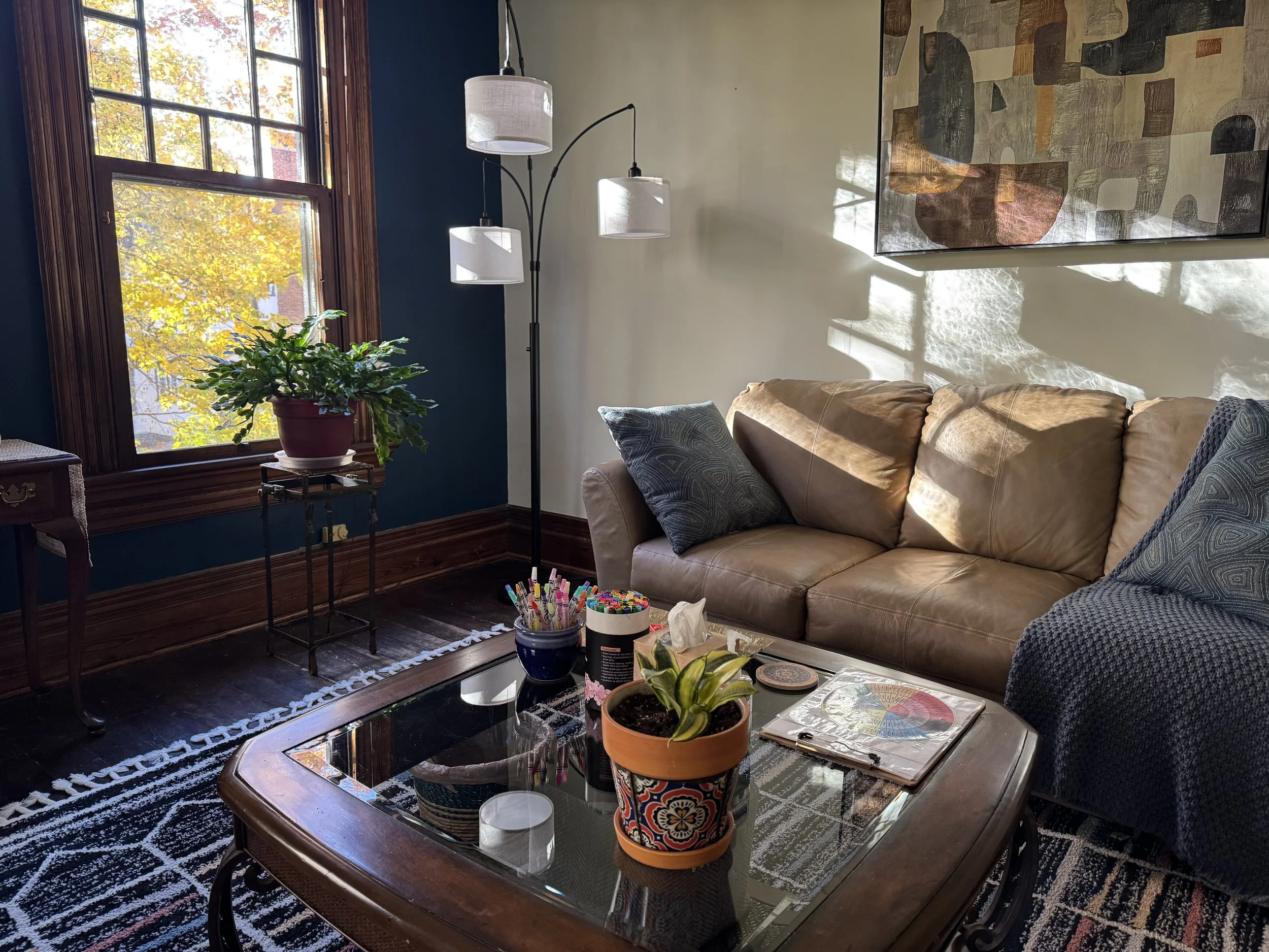 Living room with a beige sofa, dark patterned throw pillows, wooden coffee table with plants and supplies, blue accent wall, large window with leaf-filled trees outside, colorful abstract artwork on the wall, a framed art piece, and a corner lamp with three white shades.