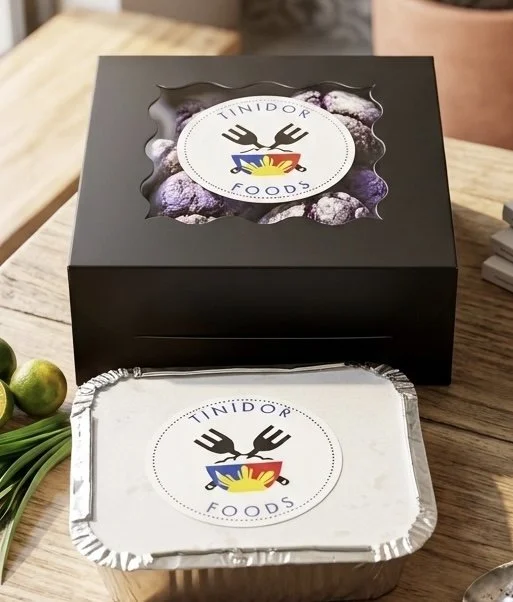 Professional Tinidor Foods catering packaging featuring a sleek black box with a window showing food inside and a foil container, both labeled with the company logo