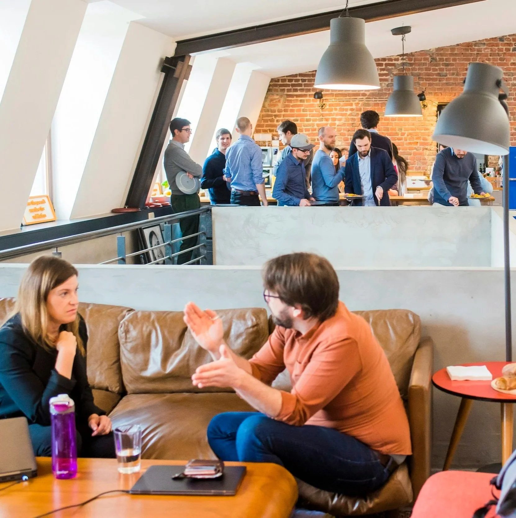 People in a busy, modern office lounge, with some engaged in conversation on a leather sofa and others standing in line at a buffet area with brick and white walls and large pendant lamps.