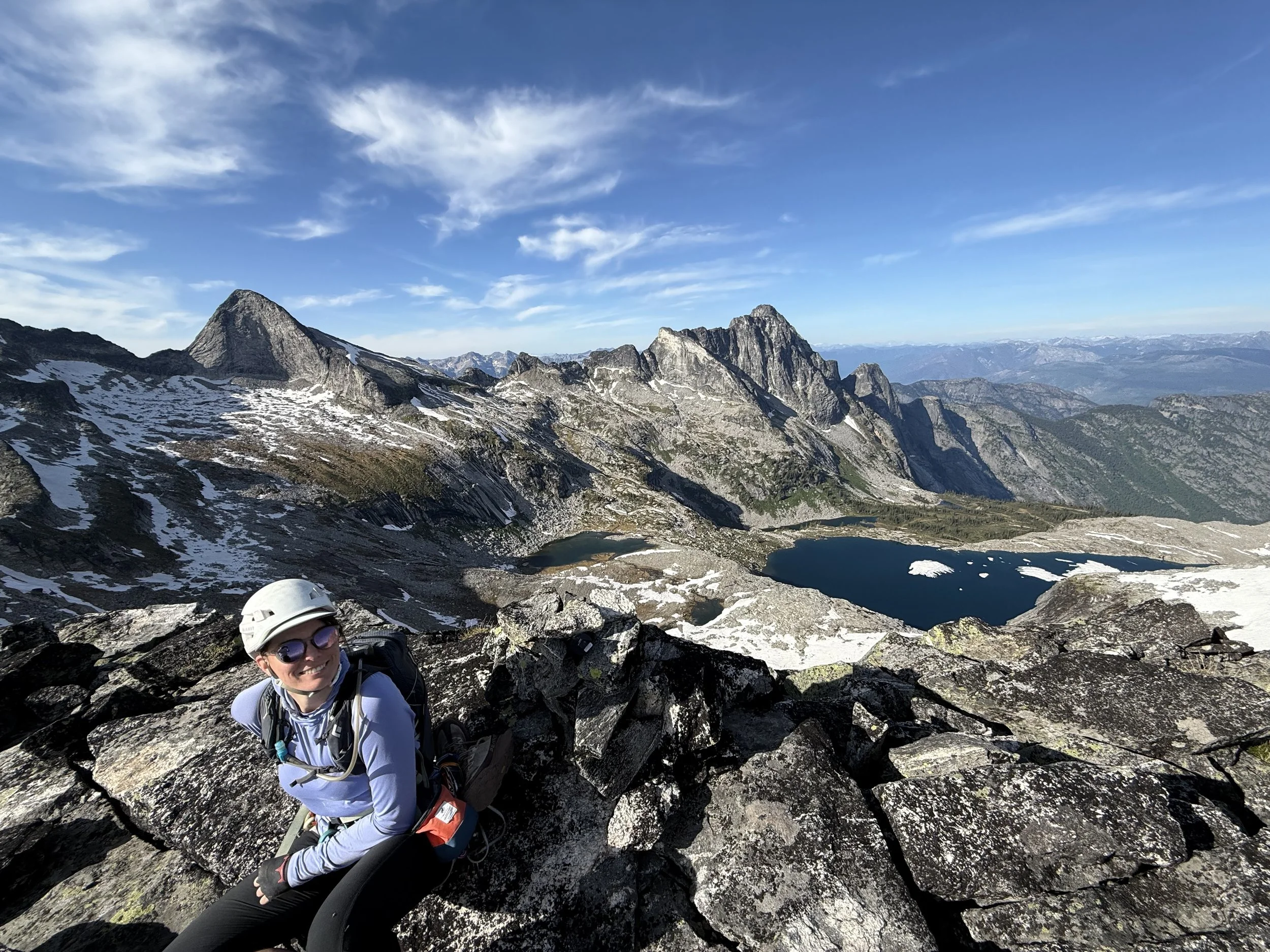 A woman wearing sunglasses and a helmet sitting on a rocky mountain terrain. She is dressed in outdoor gear and smiling. Behind her, rugged mountains with patches of snow and a small lake are visible under a blue sky with scattered clouds.