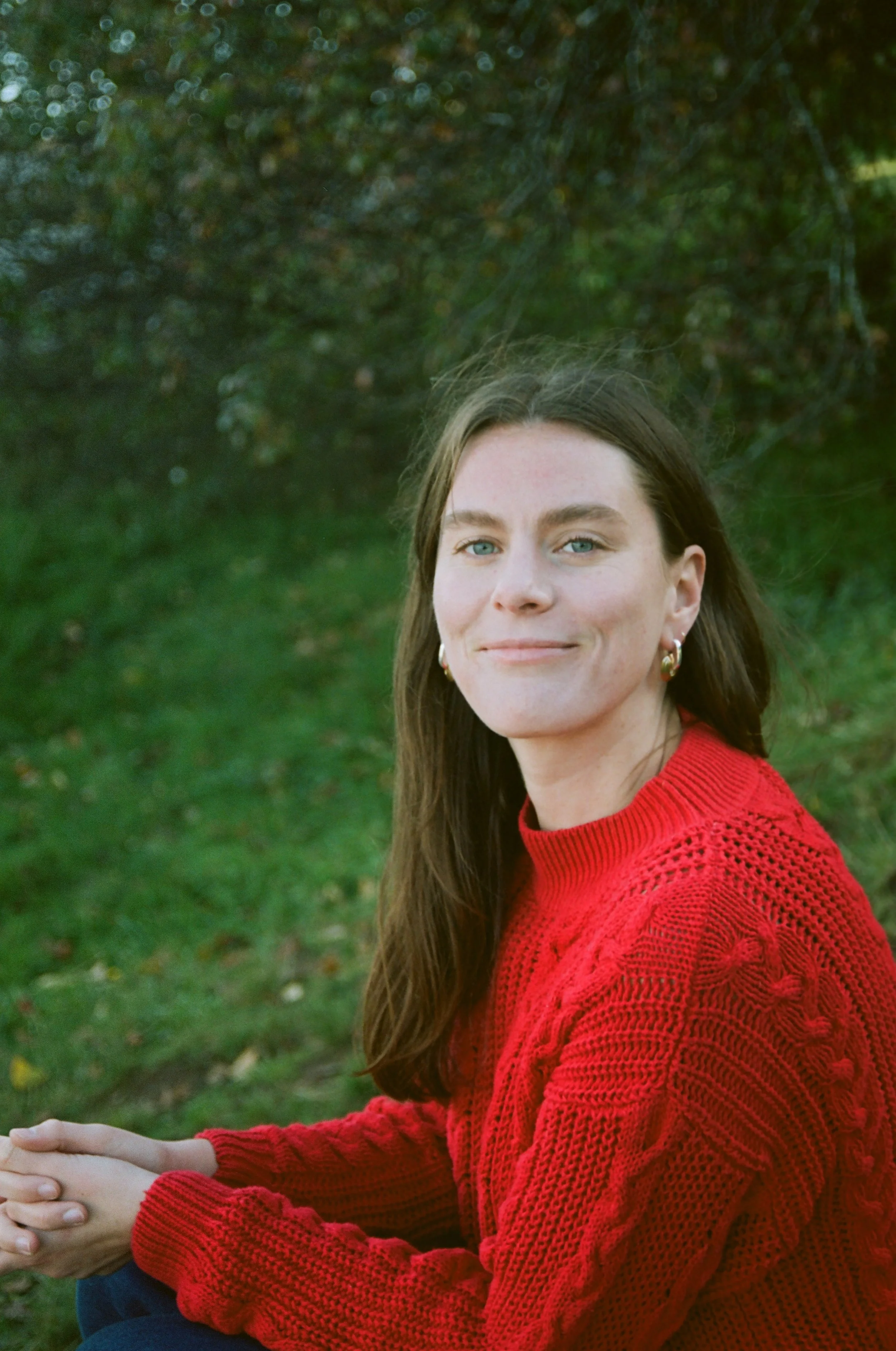 A woman with long brown hair and blue eyes, wearing a red knitted sweater and earrings, sitting outdoors in front of a blurred green background with trees.