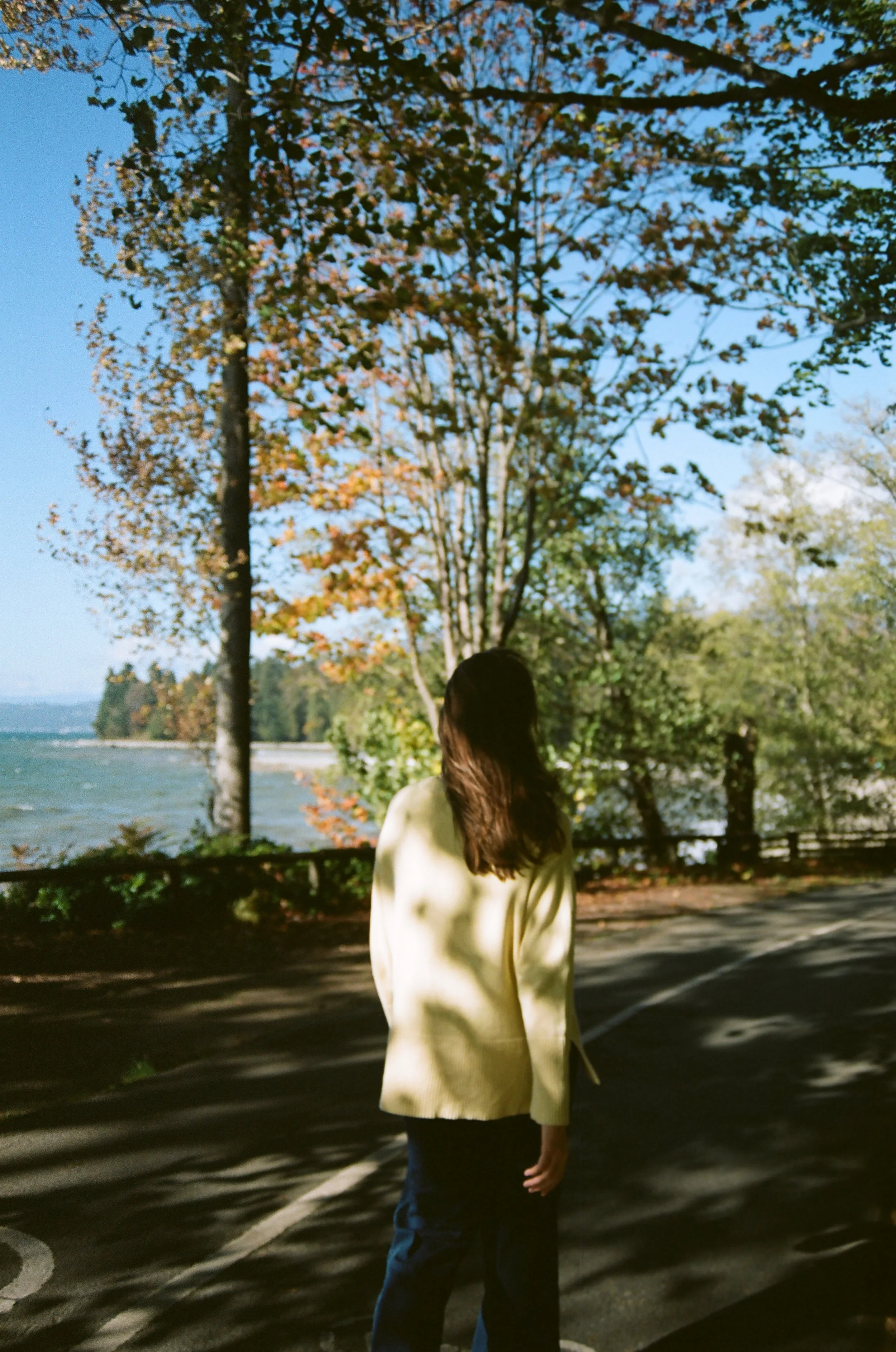 A woman with long brown hair, wearing a beige coat, standing on a paved area near a body of water, with trees and blue sky in the background.