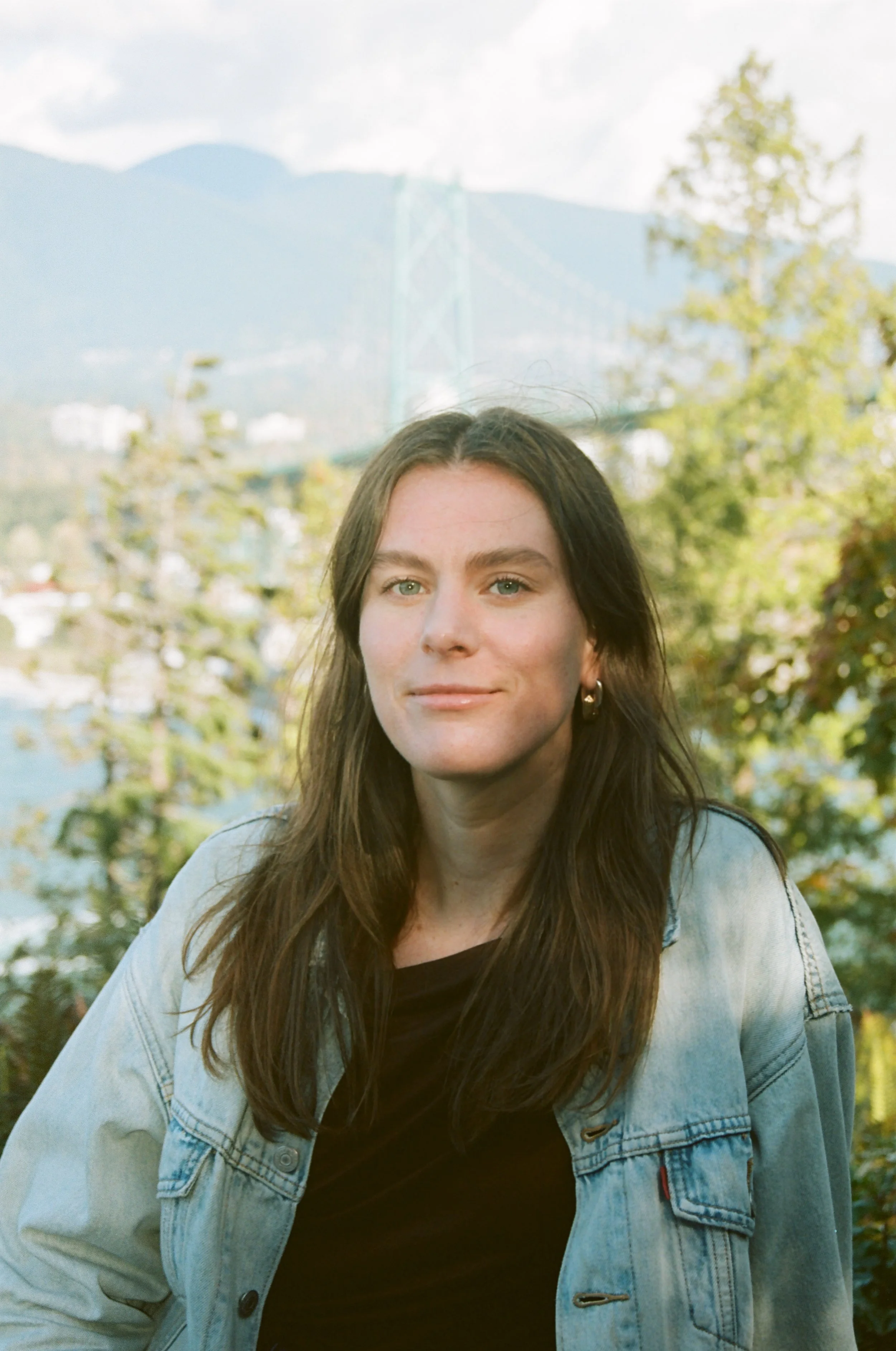 A woman with long brown hair, wearing a light denim jacket over a black top, sitting outdoors with trees and mountains in the background.