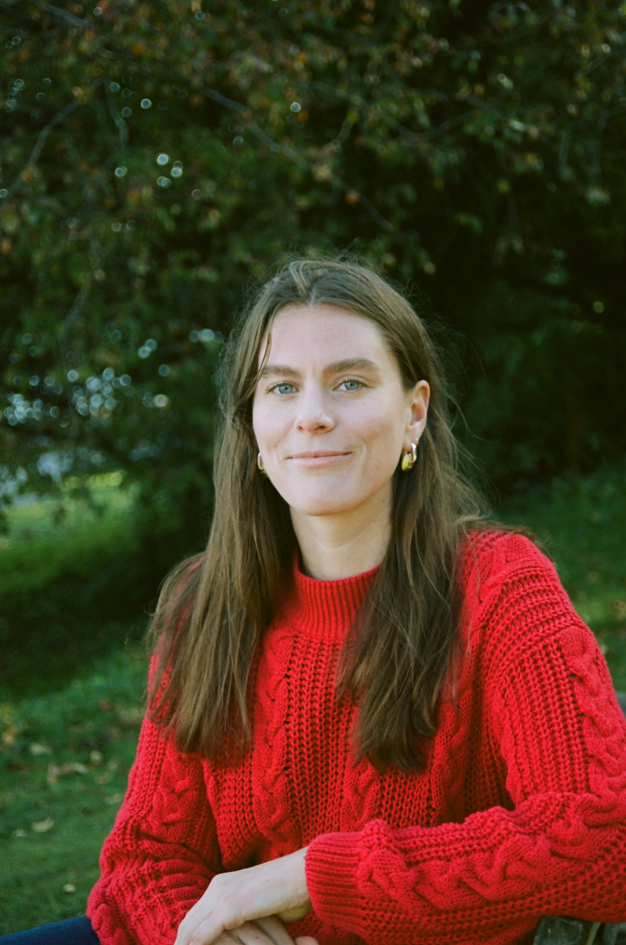 A woman with long brown hair, blue eyes, and hoop earrings, wearing a red cable-knit sweater, outdoors with greenery and trees in the background.
