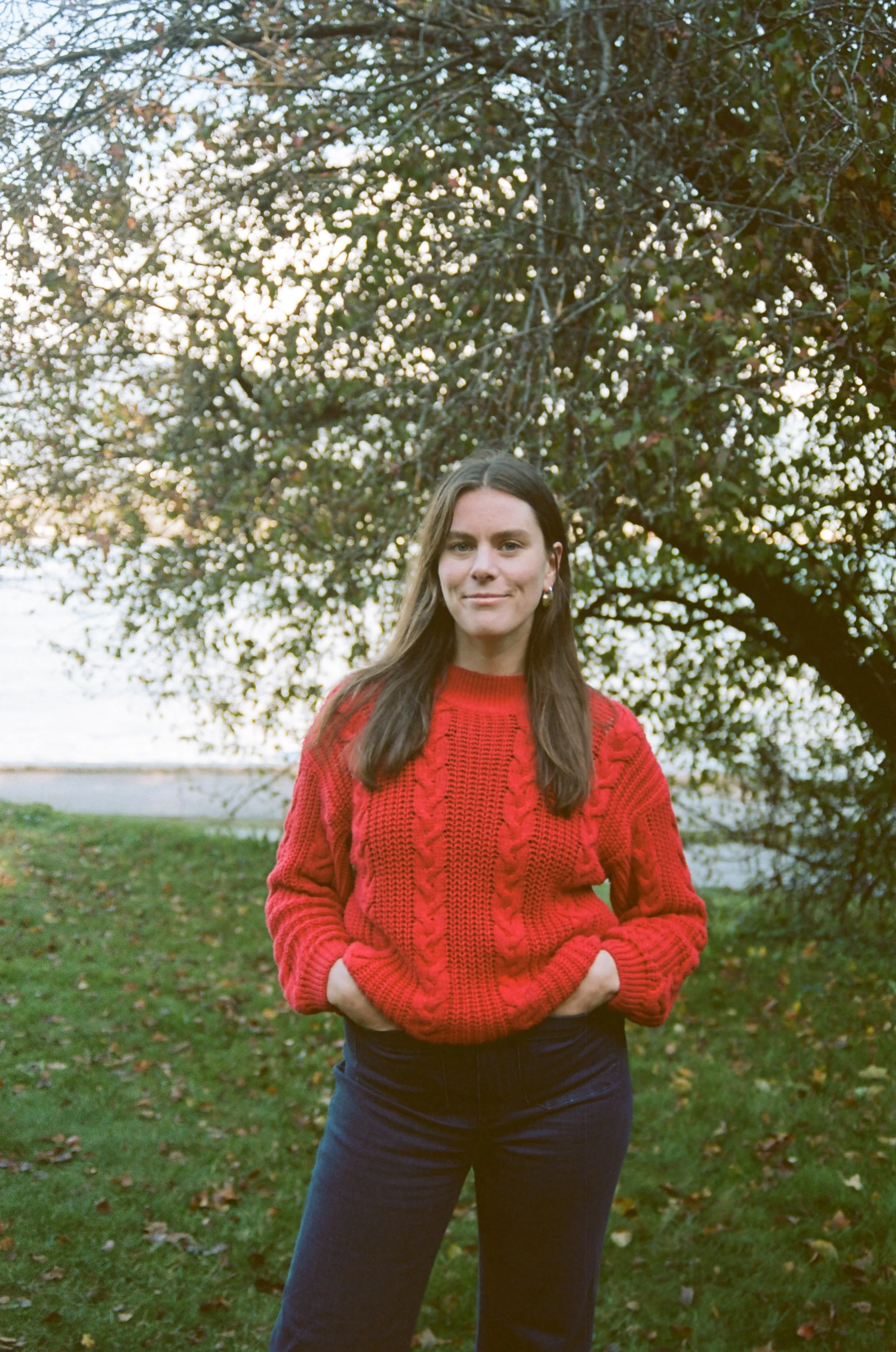 A woman standing outdoors in front of a leafy tree, wearing a red sweater and dark pants, with her hands in her pockets.