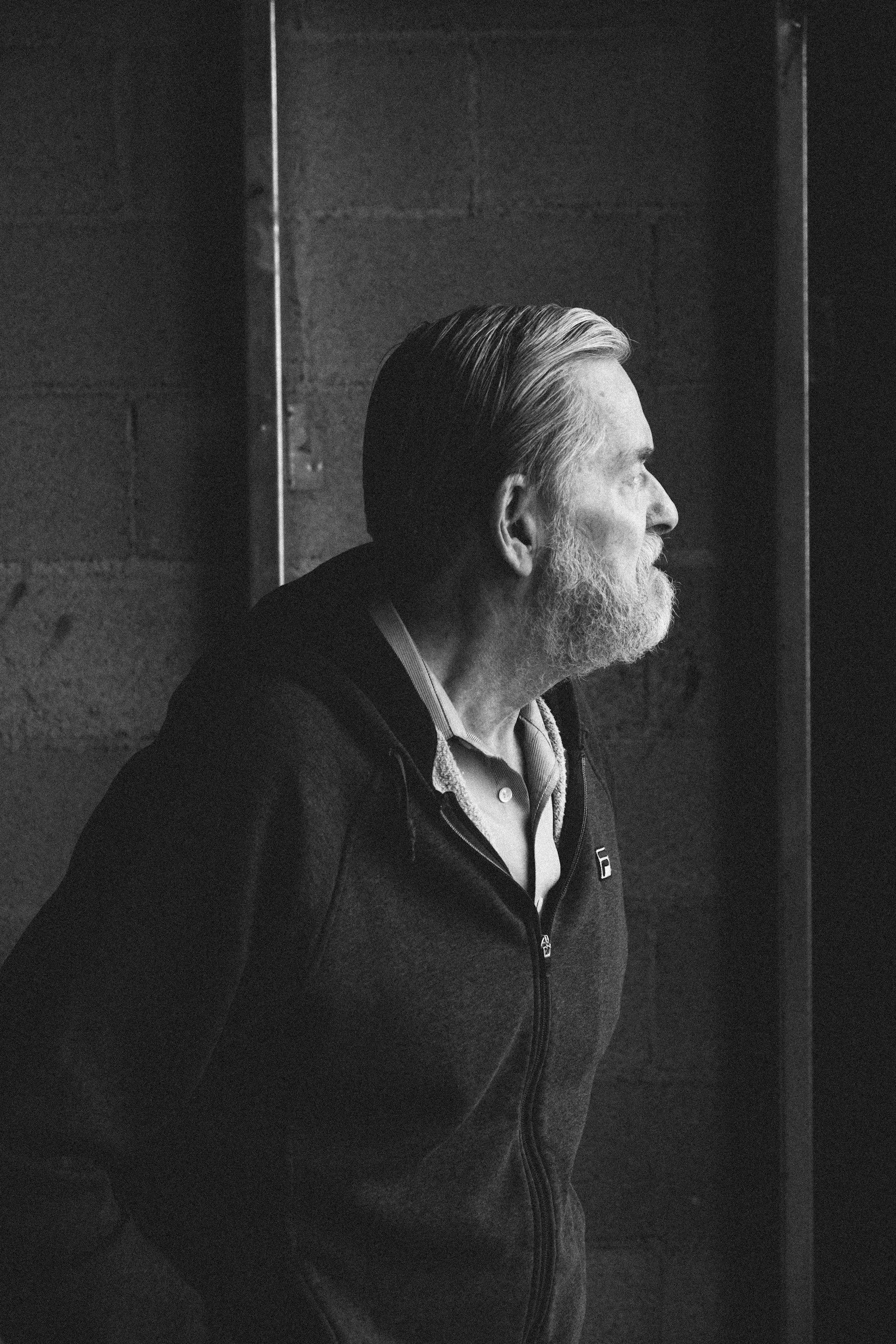 Side profile of a mature man with gray hair and beard, wearing a dark zip-up jacket, standing against a textured wall in black and white.