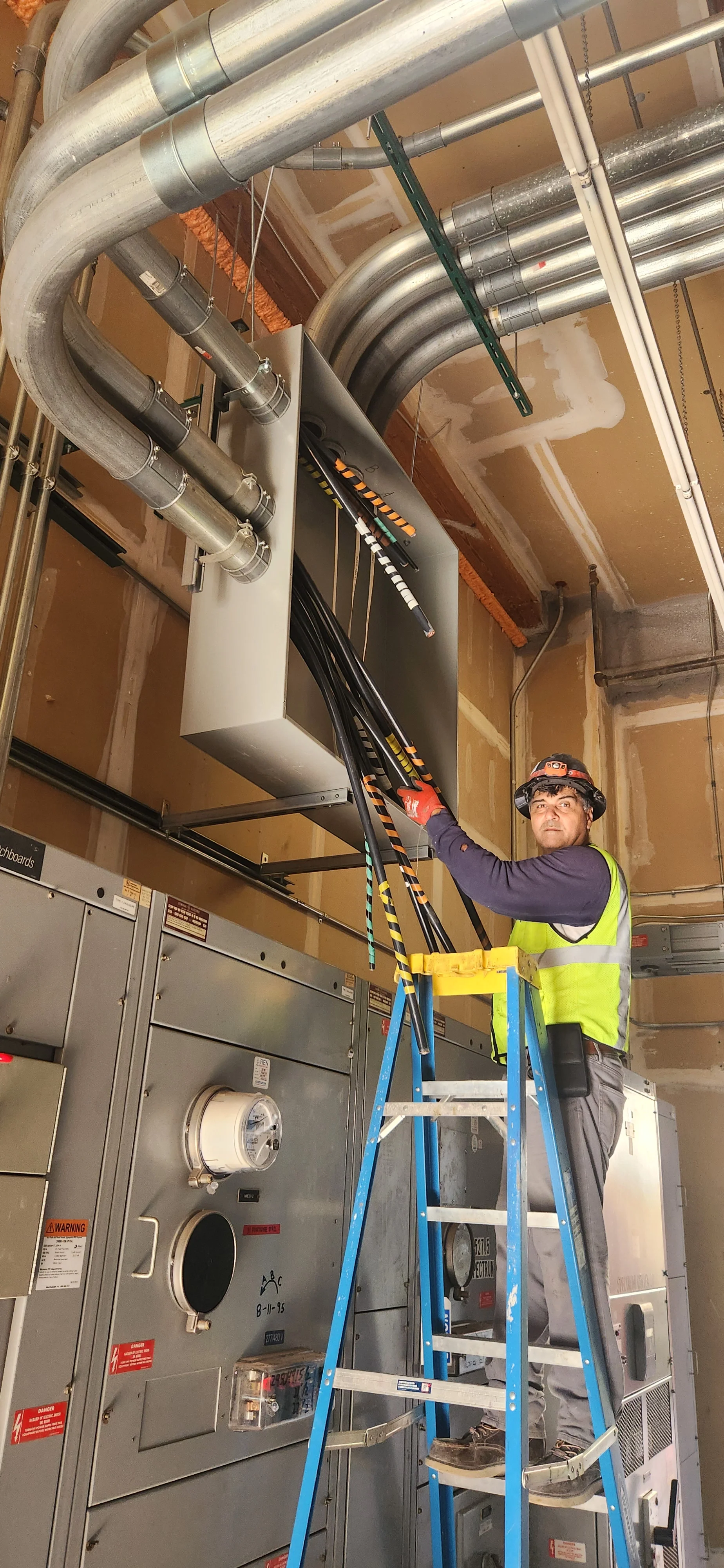 A worker wearing a safety vest, helmet, and gloves is standing on a ladder, working on electrical or HVAC equipment in an industrial setting with exposed ductwork and electrical panels.