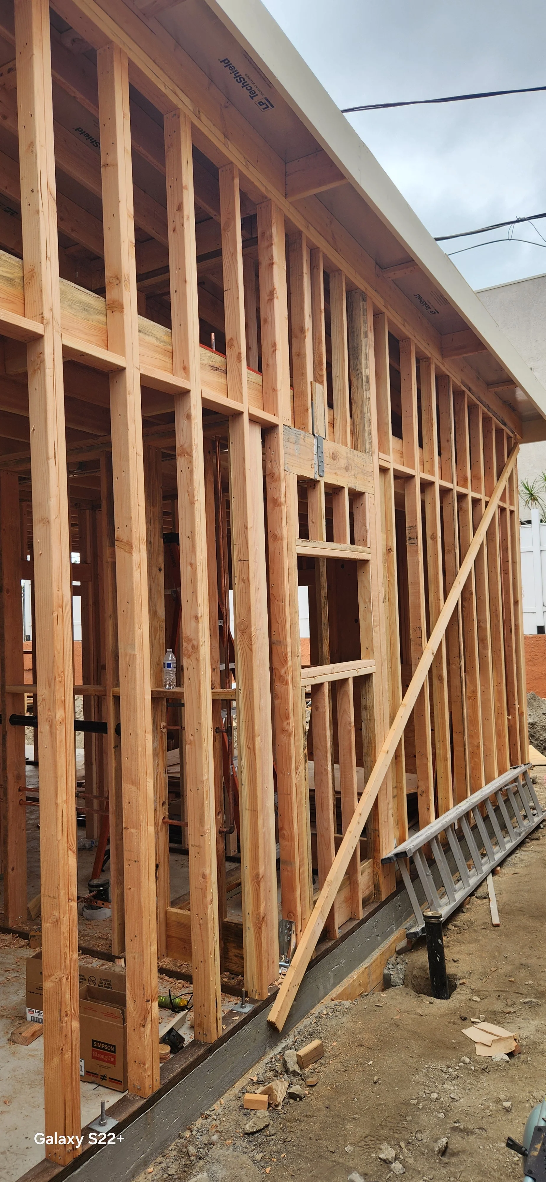 Construction site showing the wooden framing of a building with a concrete foundation and a sloped roof, with tools and supplies visible around.