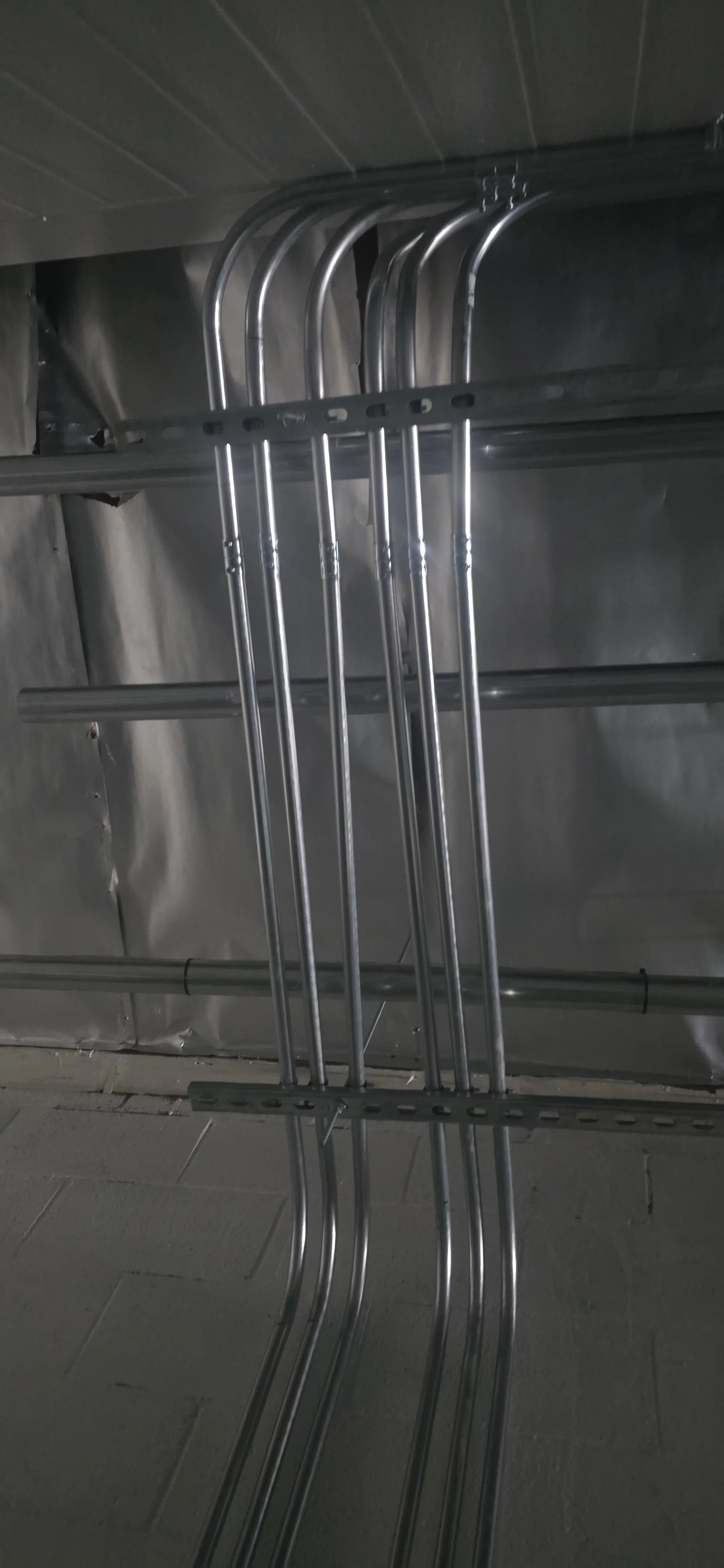 Metal pipes and electrical conduit arranged on the floor and mounted on a wall in an industrial setting.