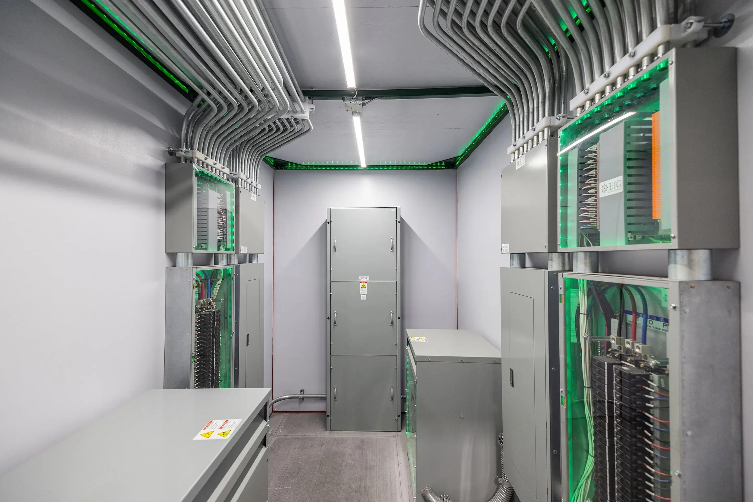 A data center with gray electrical equipment and numerous network cables organized along the top and sides, illuminated with green LED lights.