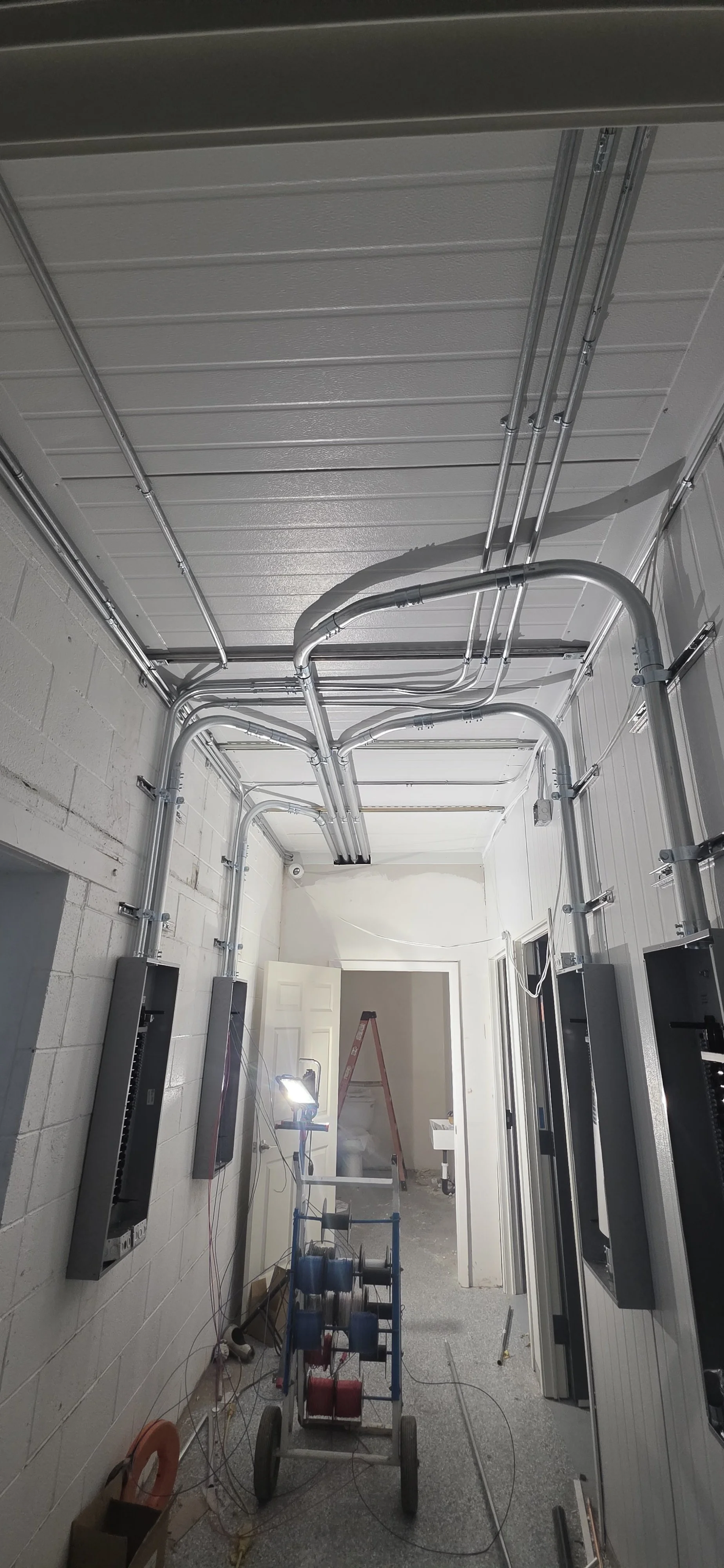 Construction room with installed electrical conduits on ceiling and walls, ladder, work light, and electrical wiring.