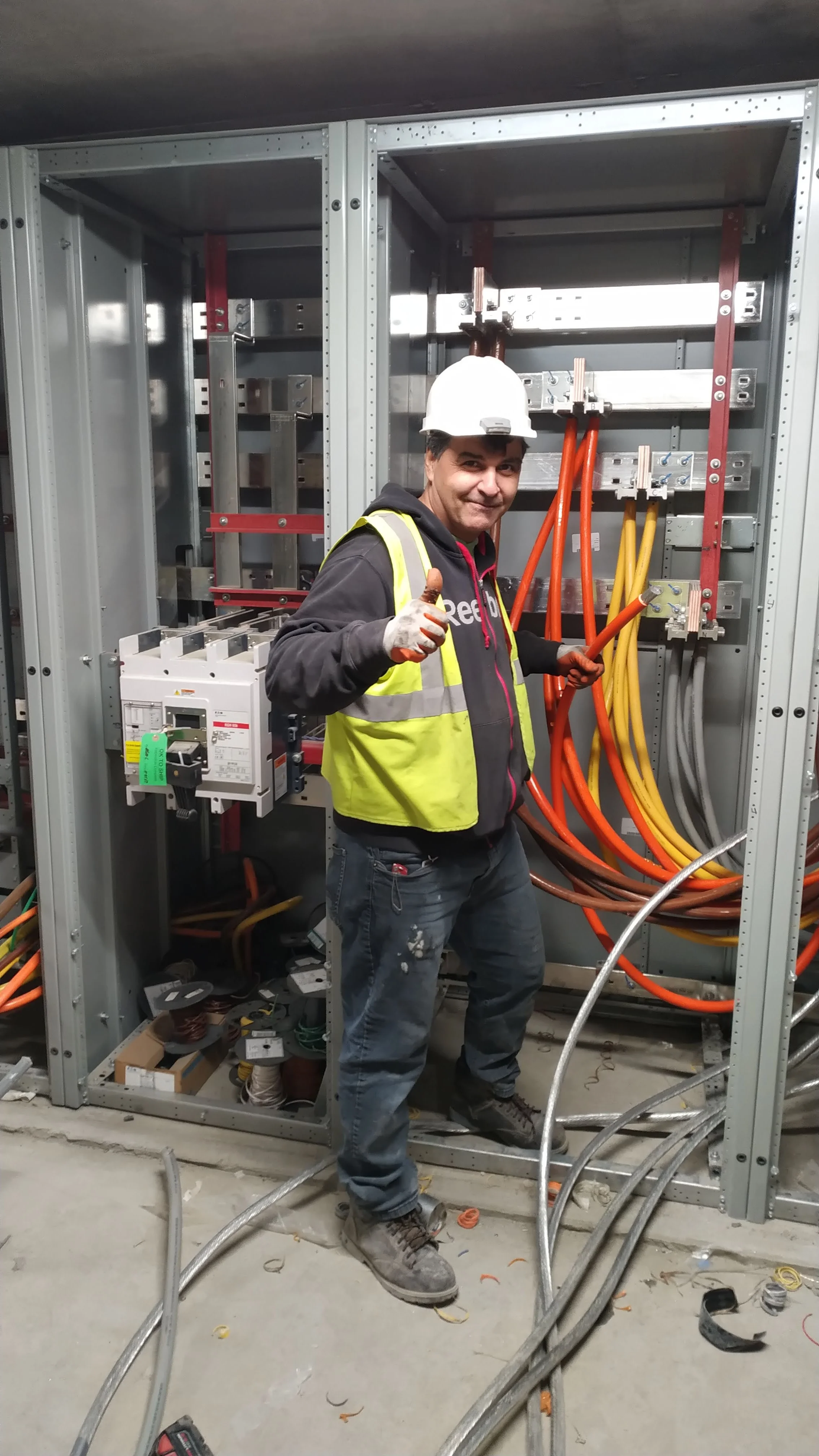 A construction worker wearing a white hard hat, yellow safety vest, and gloves, standing inside an electrical room, holding orange electrical cables while giving a thumbs-up.