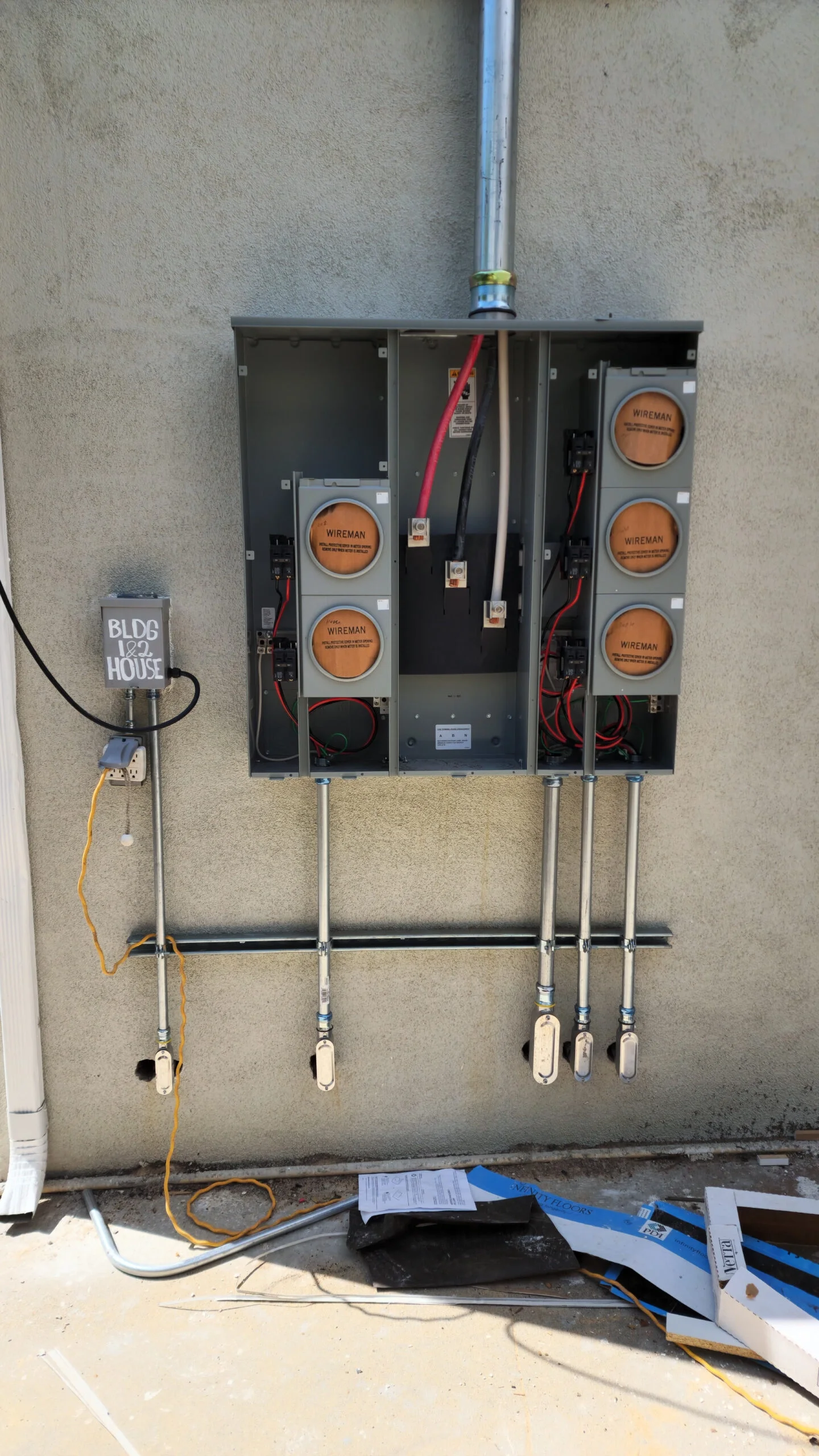 An outdoor electrical panel with open wiring and components, mounted on a wall, with a small box labeled 'BLDG 162 HOUSE' attached to a conduit, and various tools and materials on the ground below.