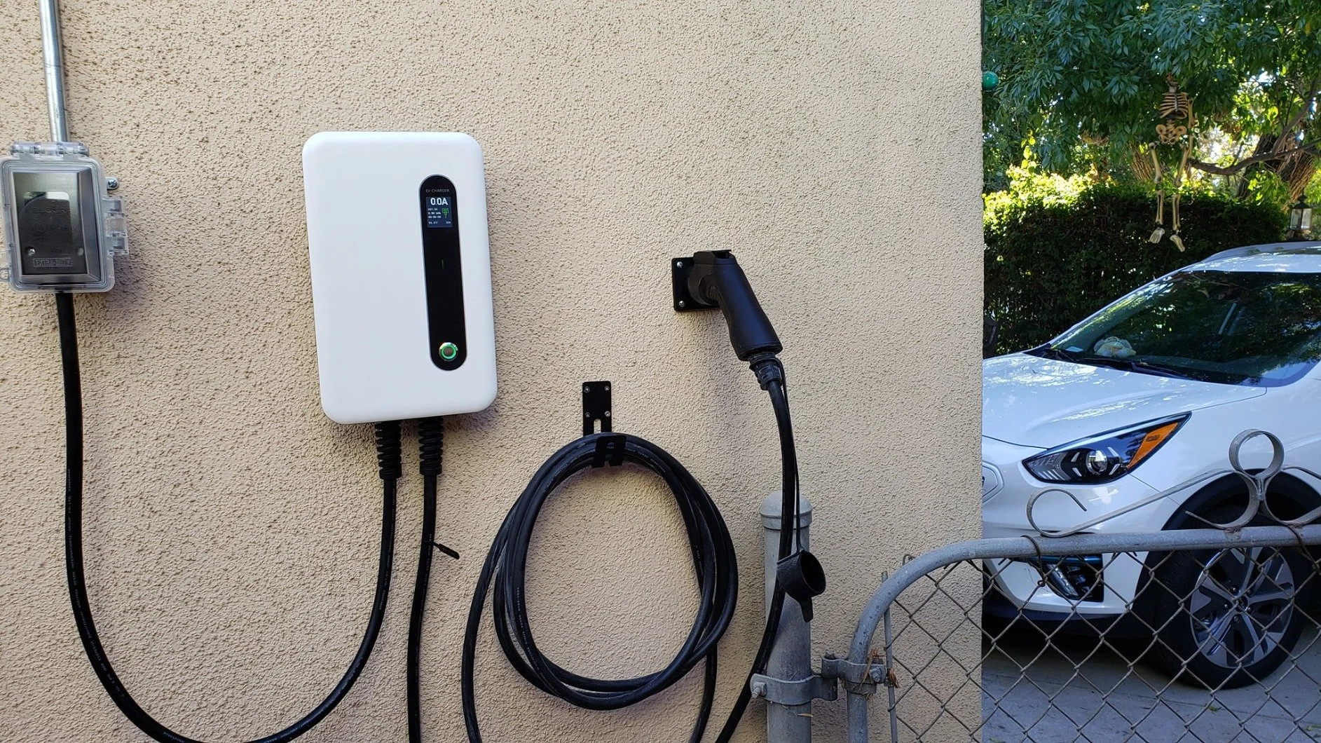 Electric vehicle charging station on a beige exterior wall, with a charging cable, next to a white car in a residential driveway, and a decorative skeleton hanging from a tree in the background.