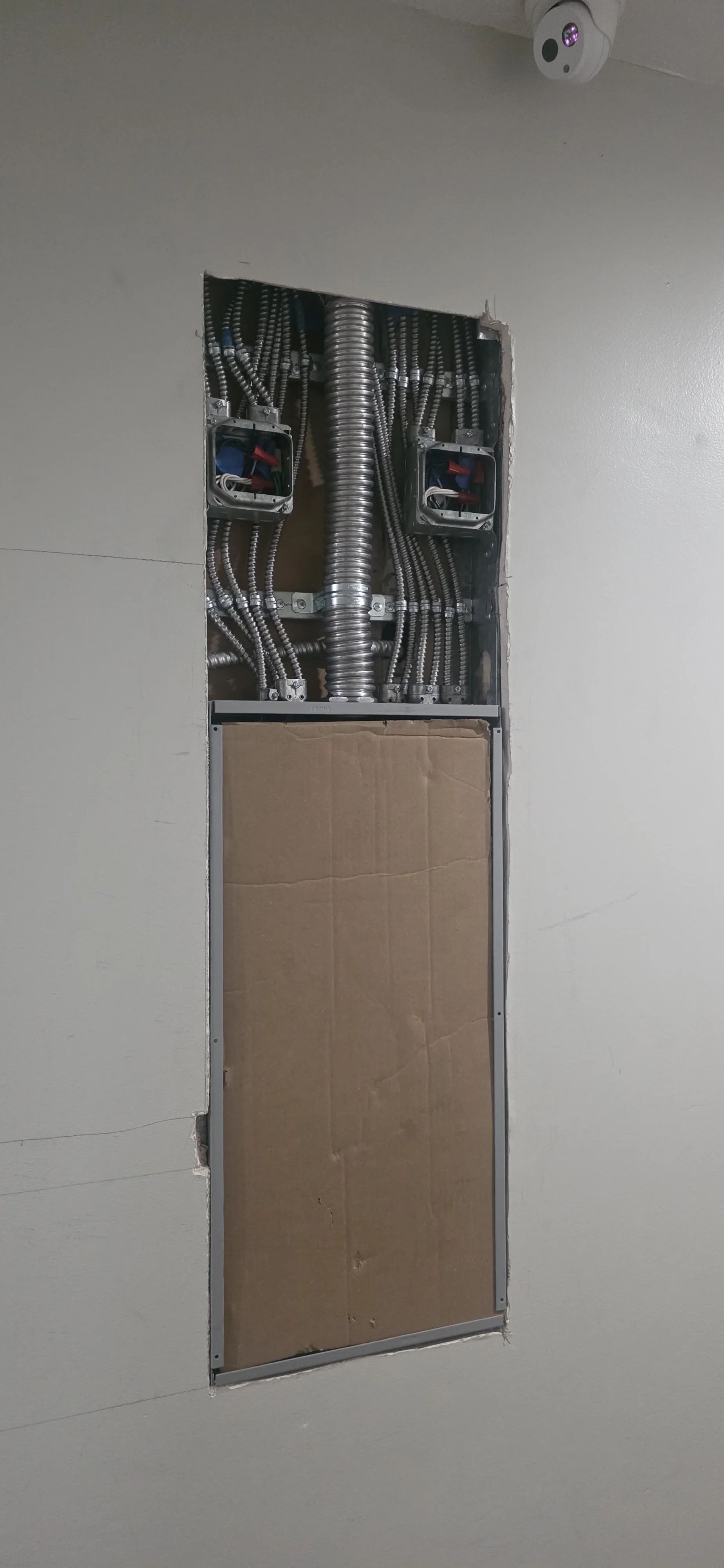 Construction wall opening with electrical wiring, metal conduit pipes, and a cardboard sheet covering part of the opening, with a security camera mounted on the ceiling.