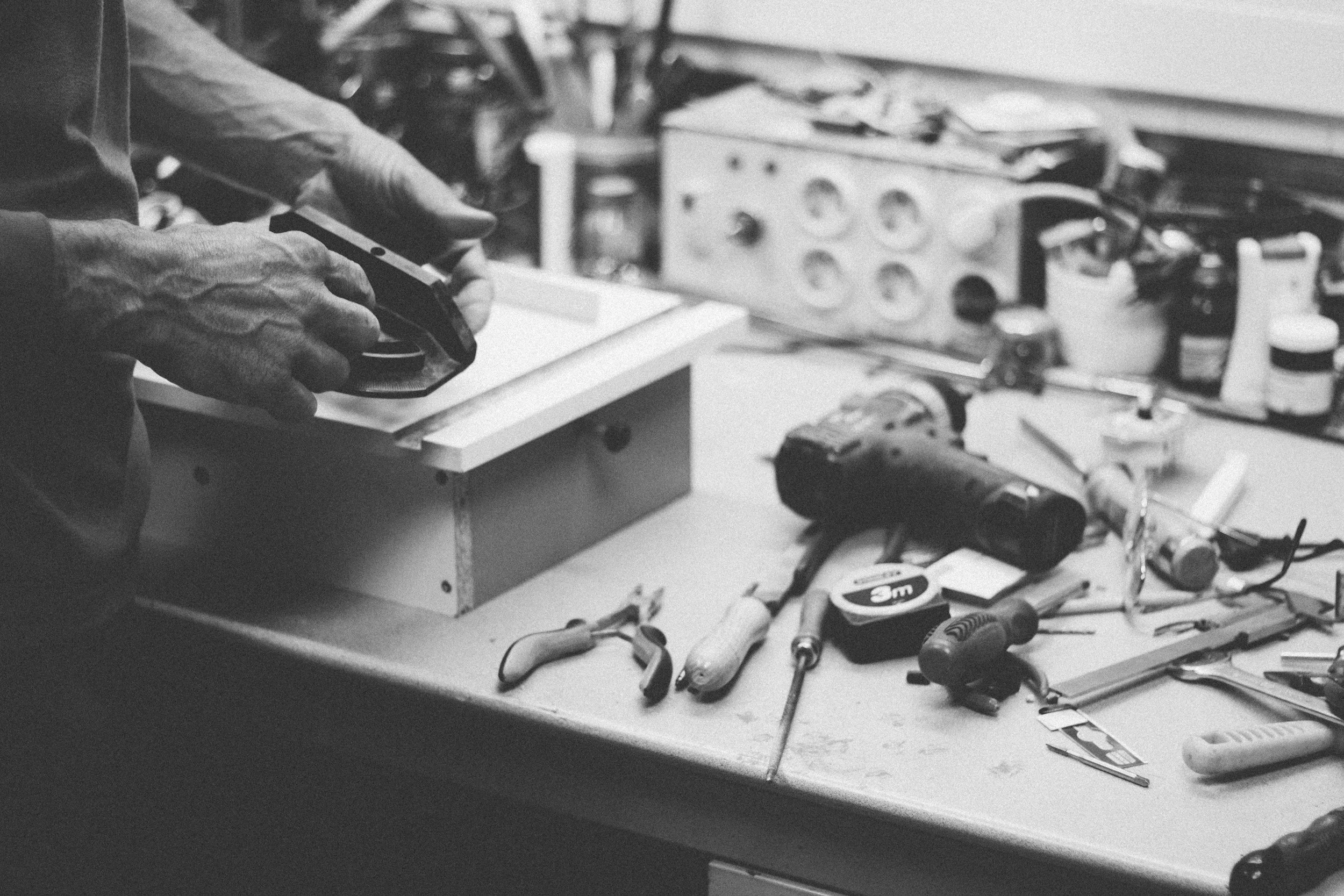 Hands holding a tool on a cluttered workshop table filled with various tools, power drill, measuring tape, and small containers.