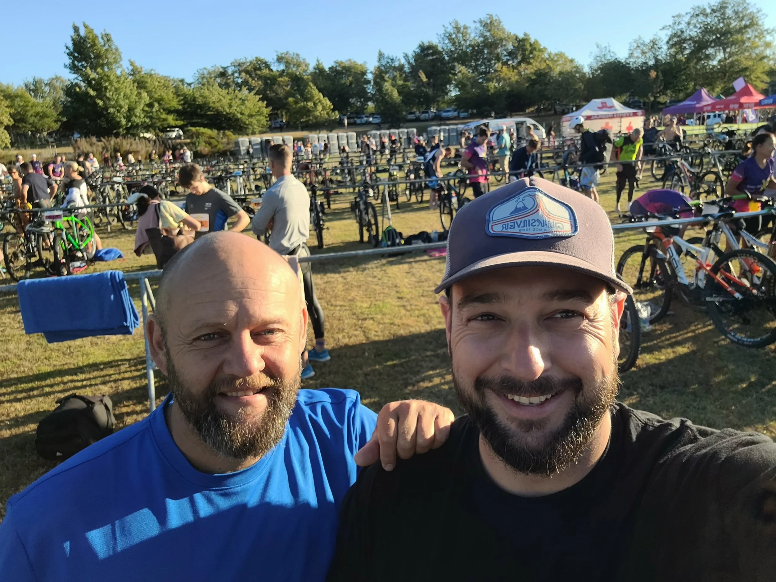 Two men smiling for a selfie at a cycling event with numerous bicycles and participants in the background.