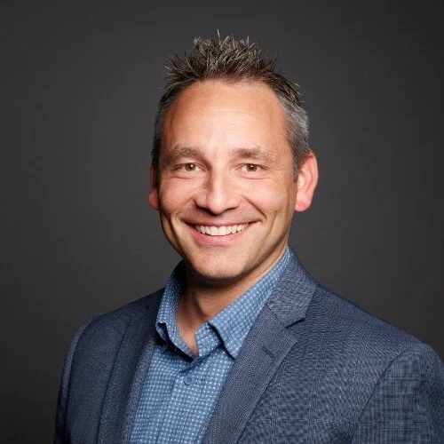 Headshot of a smiling man with short, spiked hair, wearing a blue dress shirt and blazer, against a dark background.