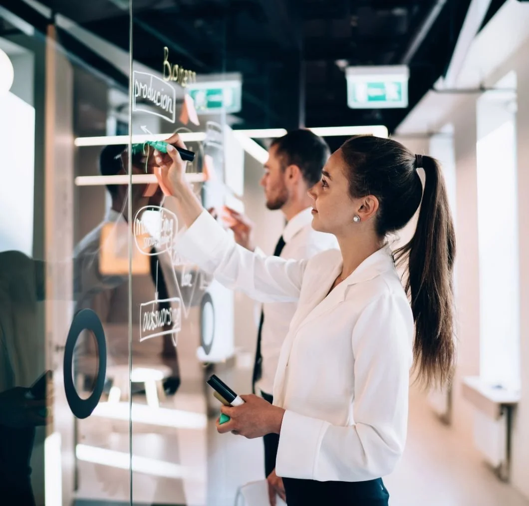 Two professionals, a woman and a man, writing on a glass board during a meeting in an office, with the woman smiling and holding a marker and a phone.