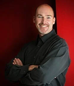 A man with a bald head and a goatee, smiling, wearing a black long-sleeve shirt, standing with arms crossed against a red background.