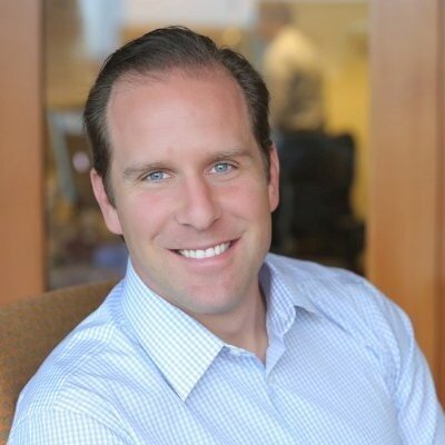 A smiling man with short, dark hair wearing a light blue dress shirt, sitting indoors with a blurred background.