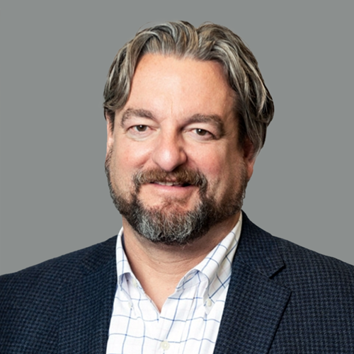Portrait of a middle-aged man with gray hair and beard, wearing a dark blazer and a white checked shirt, smiling against a gray background.