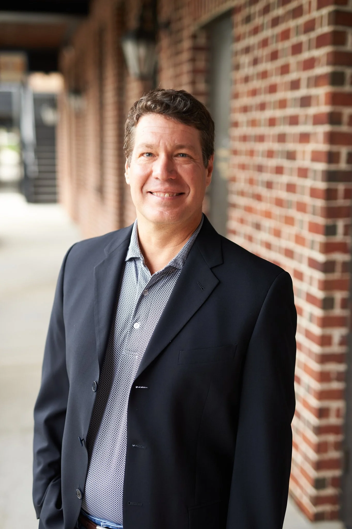 A man in a black blazer and patterned shirt standing outdoors against a brick wall and smiling.