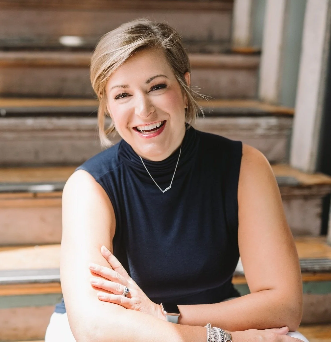 A woman with short blonde hair, smiling and wearing a black sleeveless turtleneck, sitting on wooden stairs.