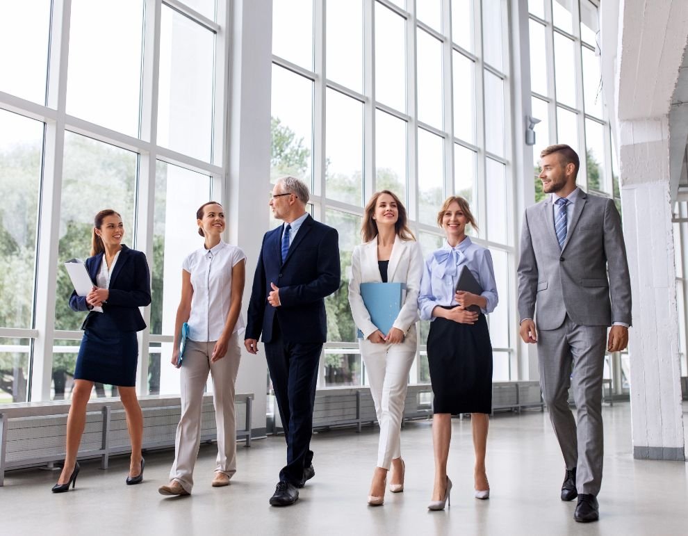 Six diverse business professionals walking together in a glass-walled lobby, smiling and holding documents.